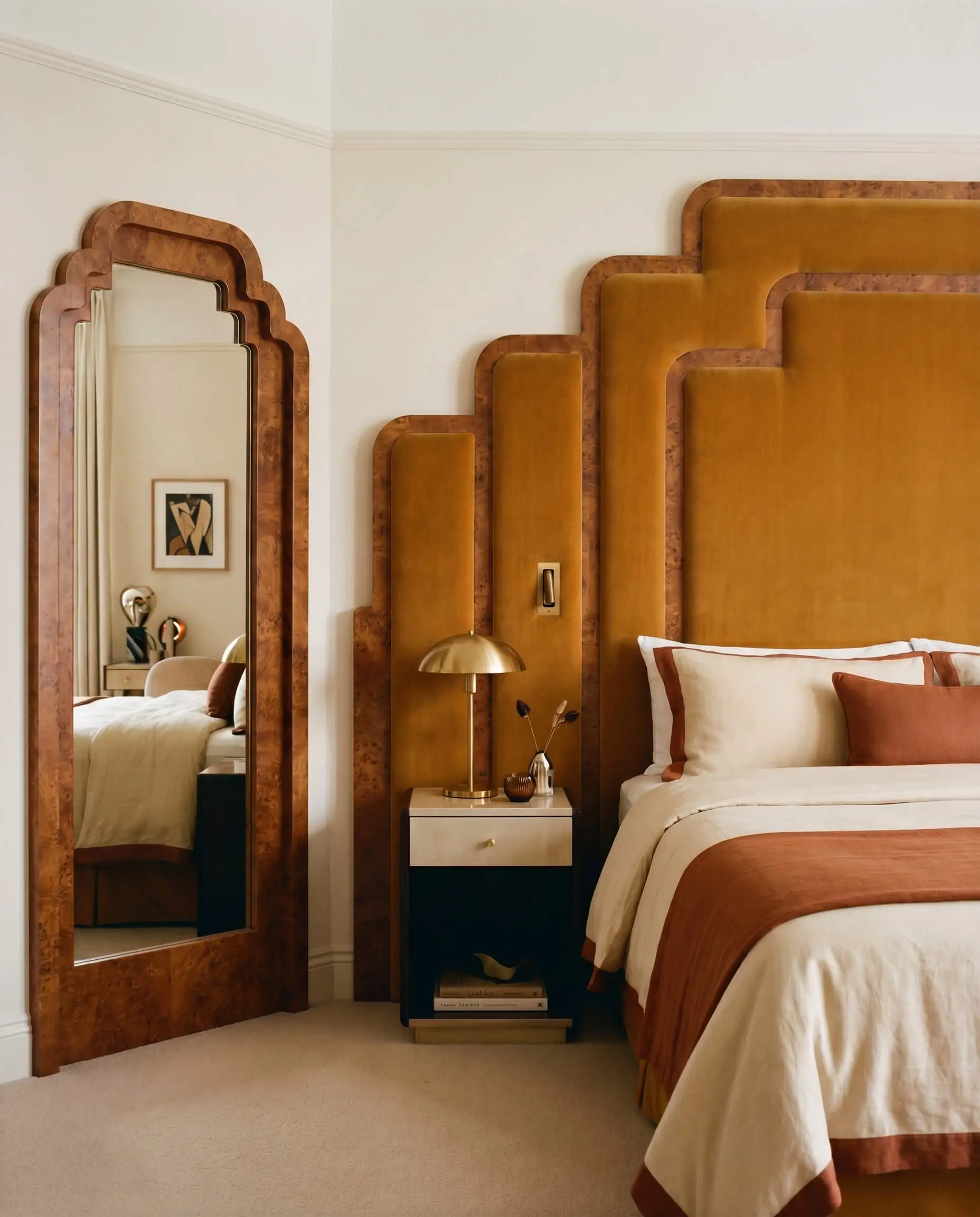 A custom stepped-arch bed surround upholstered in plush mohair with burl wood borders sits adjacent to a matching geometric vanity mirror.