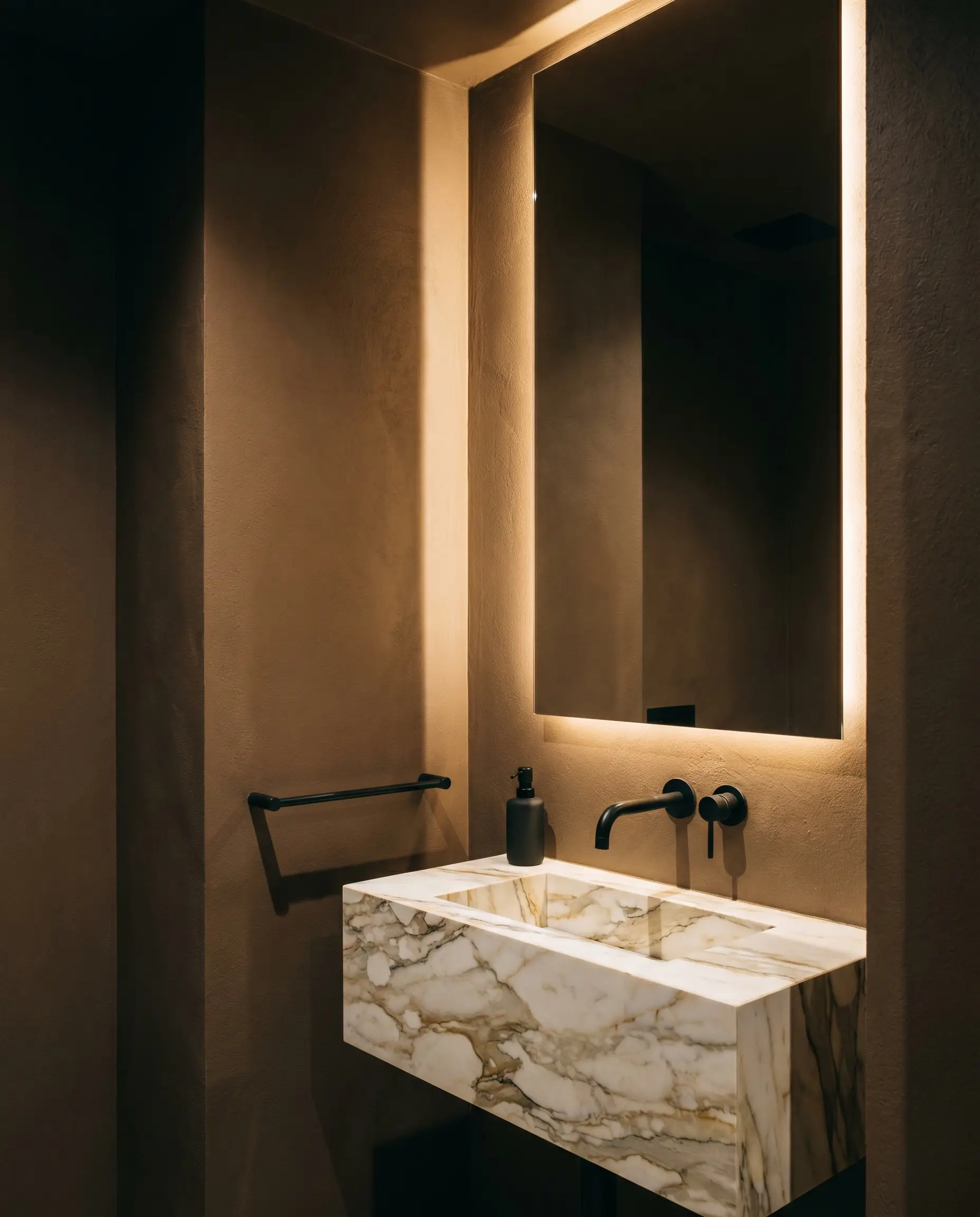 An illuminated modern mirror hangs above a striking marble vanity and sleek matte black faucet in a dramatic powder room enveloped in warm brown-beige paint.