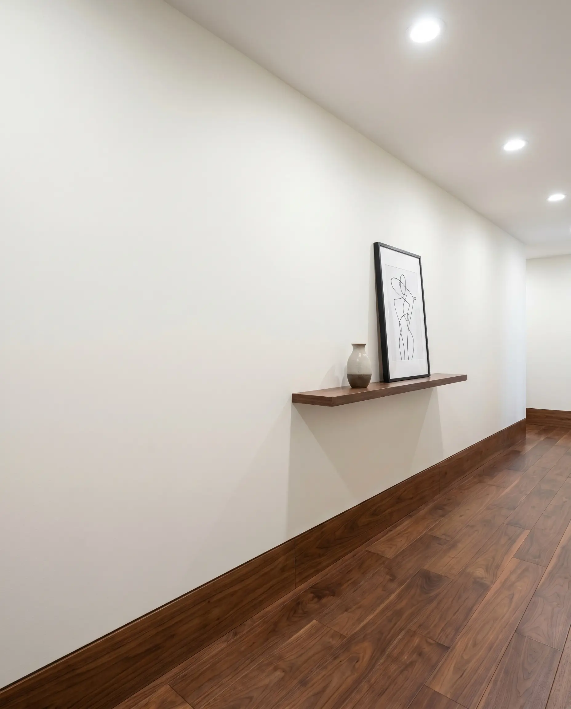 A true trimless flush baseboard system integrates a warm walnut hardwood insert seamlessly into High Reflective White drywall within a contemporary hallway.