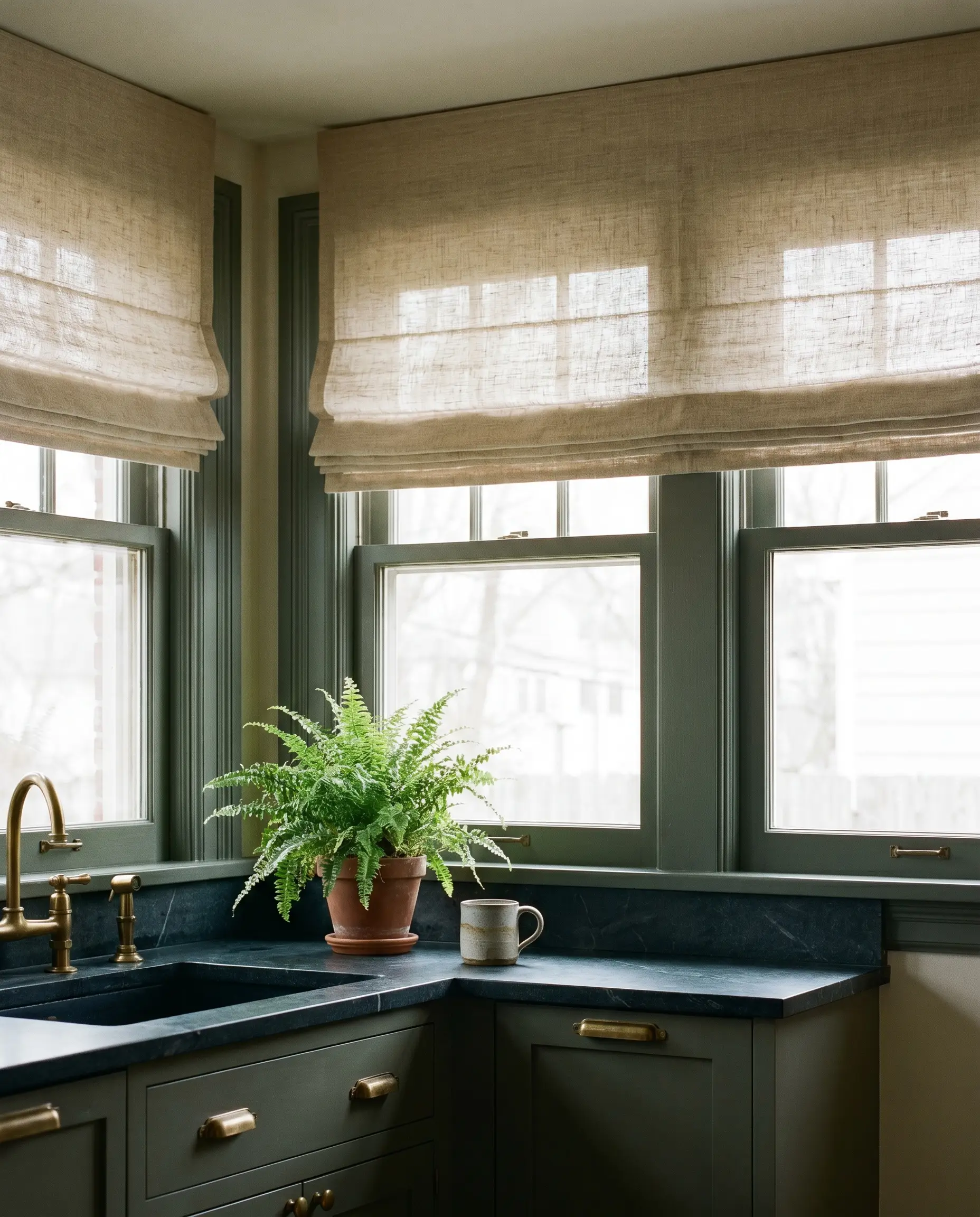 Soft unbleached linen roman shades dress a kitchen window above Farrow and Ball Green Smoke painted millwork, dark stone countertops, and brass hardware holding a potted fern.