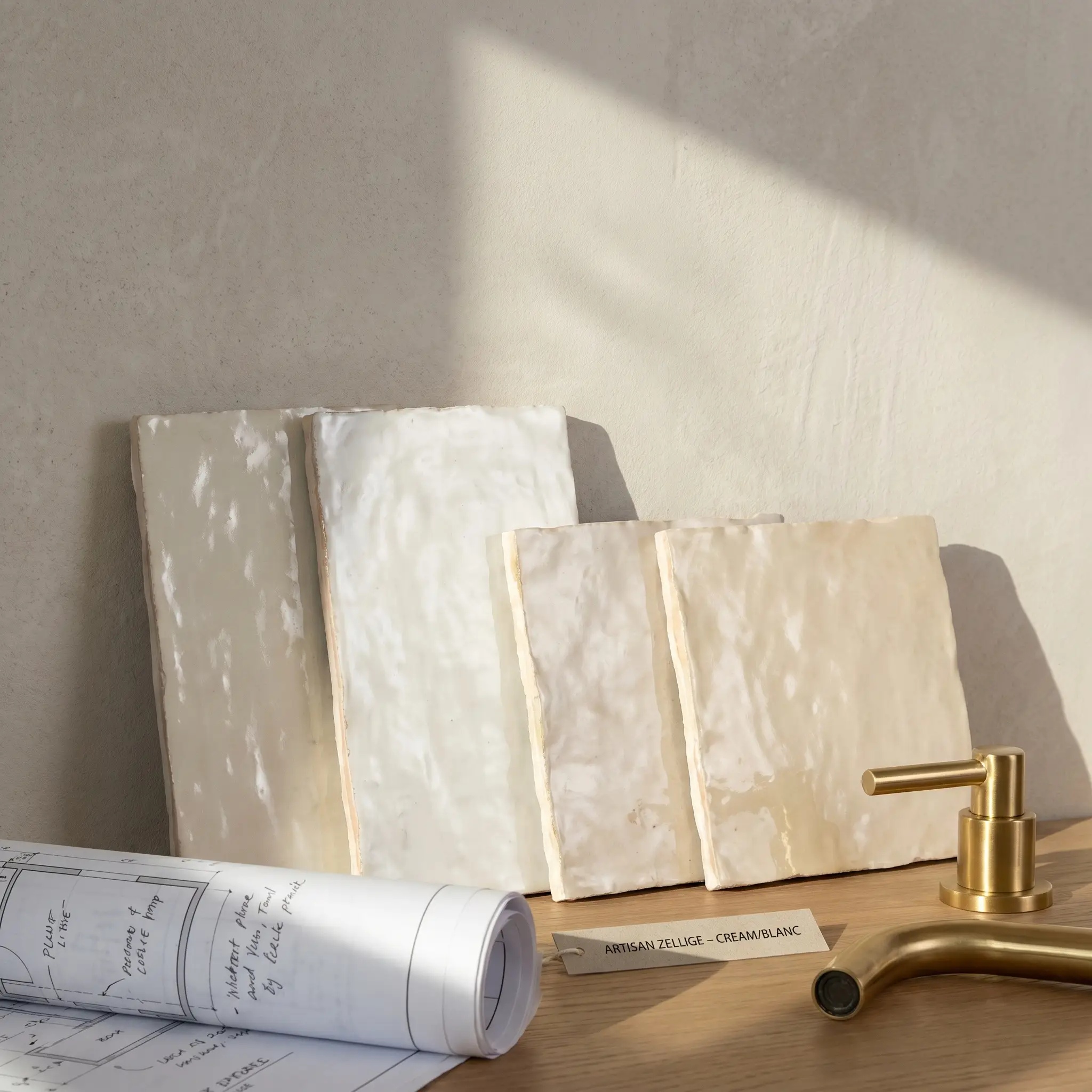 Physical sample tiles of undulating white and cream clay lean against a shower enclosure wall beside architectural blueprints and a polished brass fixture.