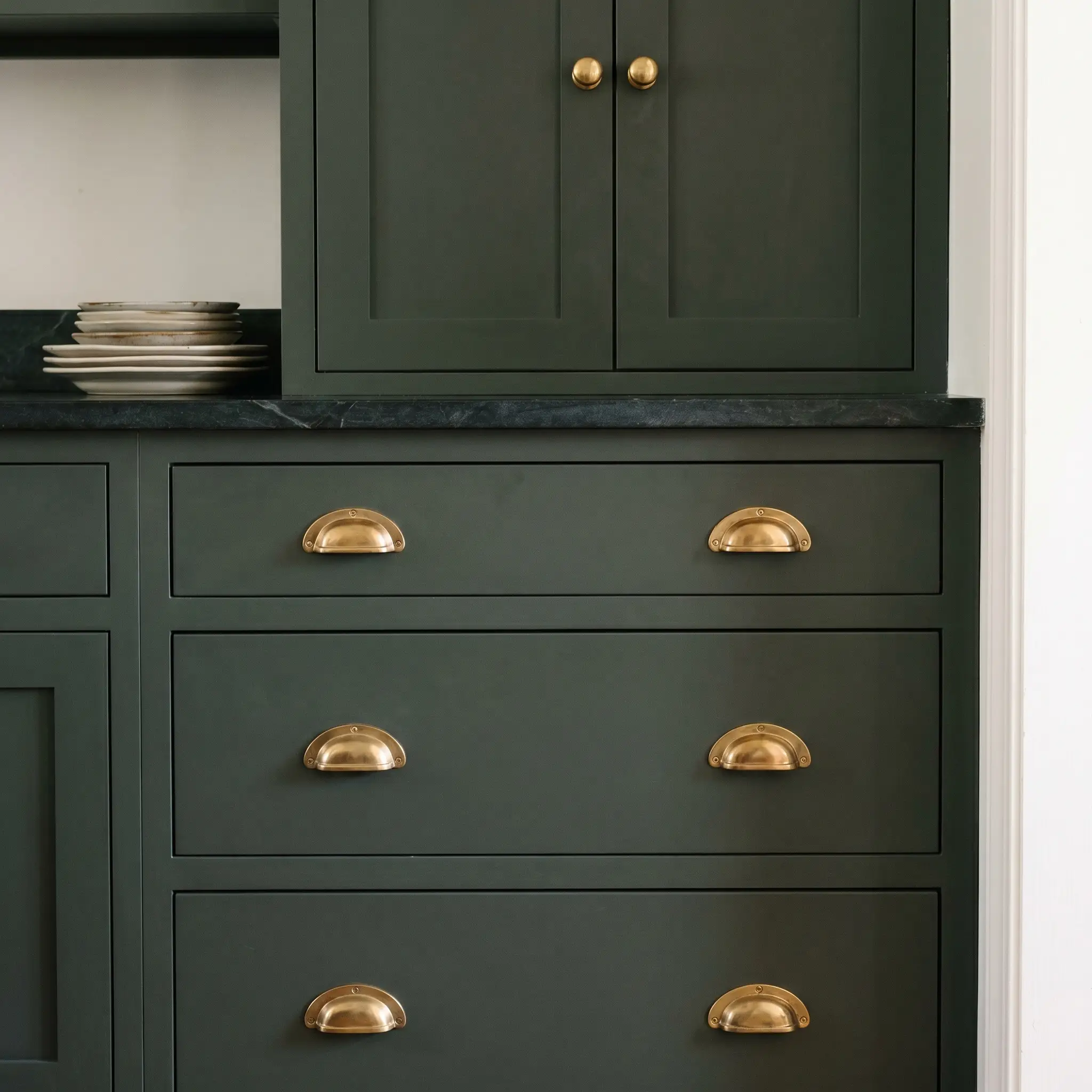 Traditional unlacquered brass cup pulls and round knobs adorn heavy Farrow and Ball Studio Green lower drawers beneath a dark steatite countertop holding heirloom plates.