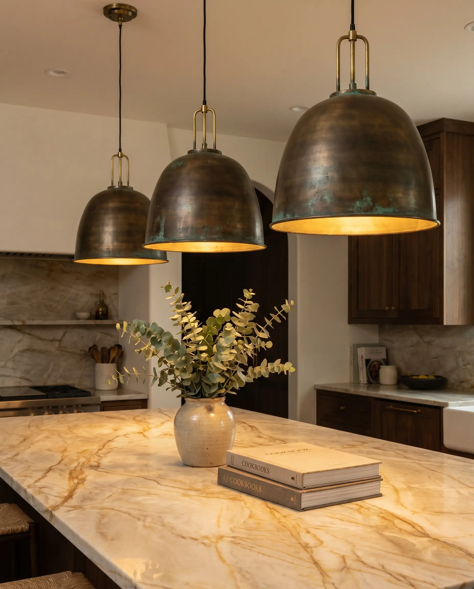 Solid unlacquered brass pendant lights cast a warm glow over Taj Mahal quartzite kitchen islands styled with greenery and luxury cookbooks.