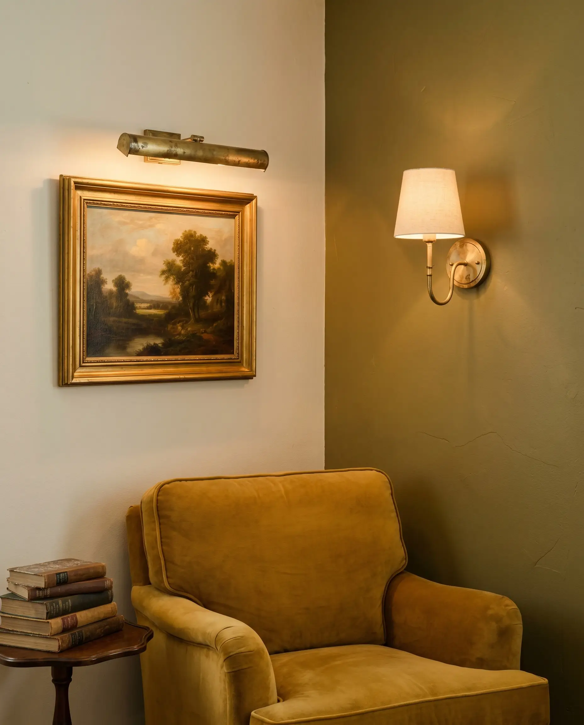 Unlacquered brass picture lights illuminate a framed landscape painting on an olive green accent wall living room featuring a mid-tier velvet armchair.
