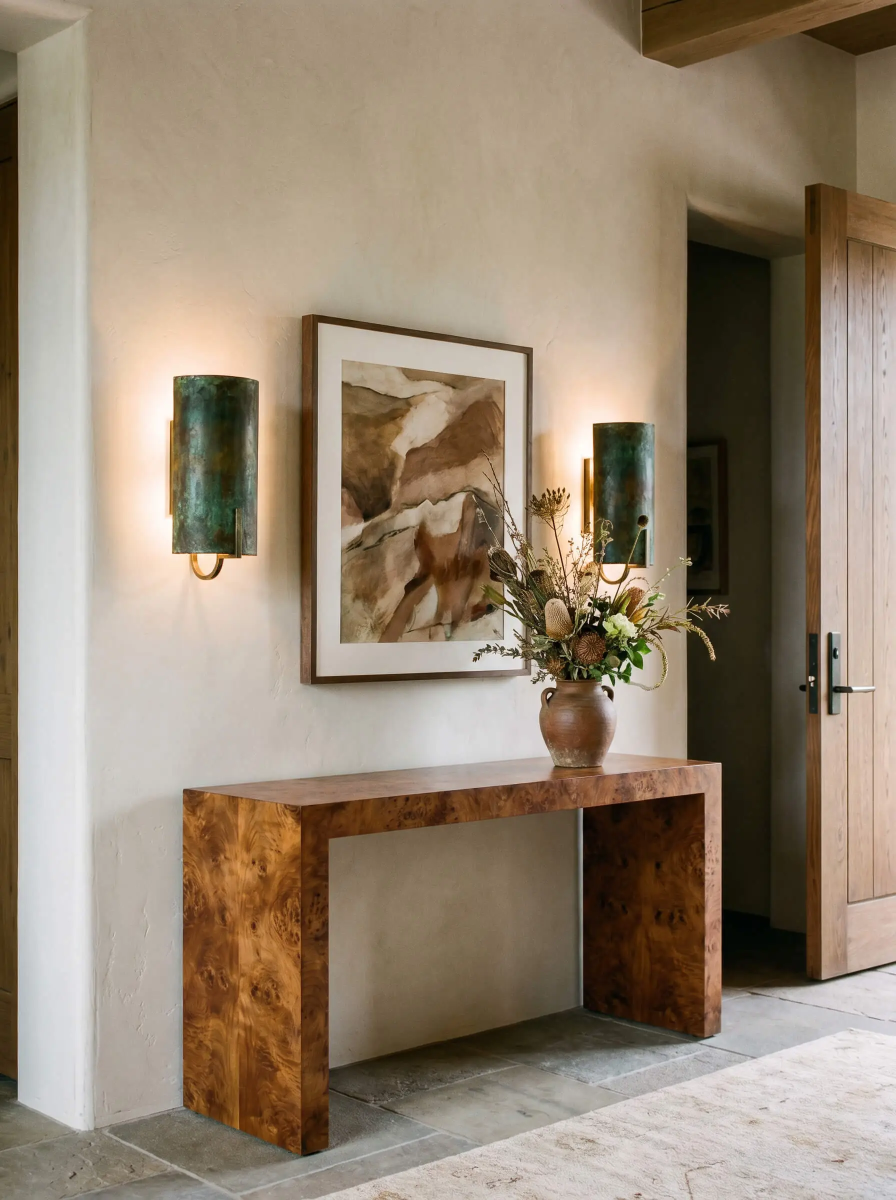 Heavy unlacquered brass and aged bronze wall sconces symmetrically flank central artwork above a warm organic burl wood console table featuring an asymmetrical floral arrangement.