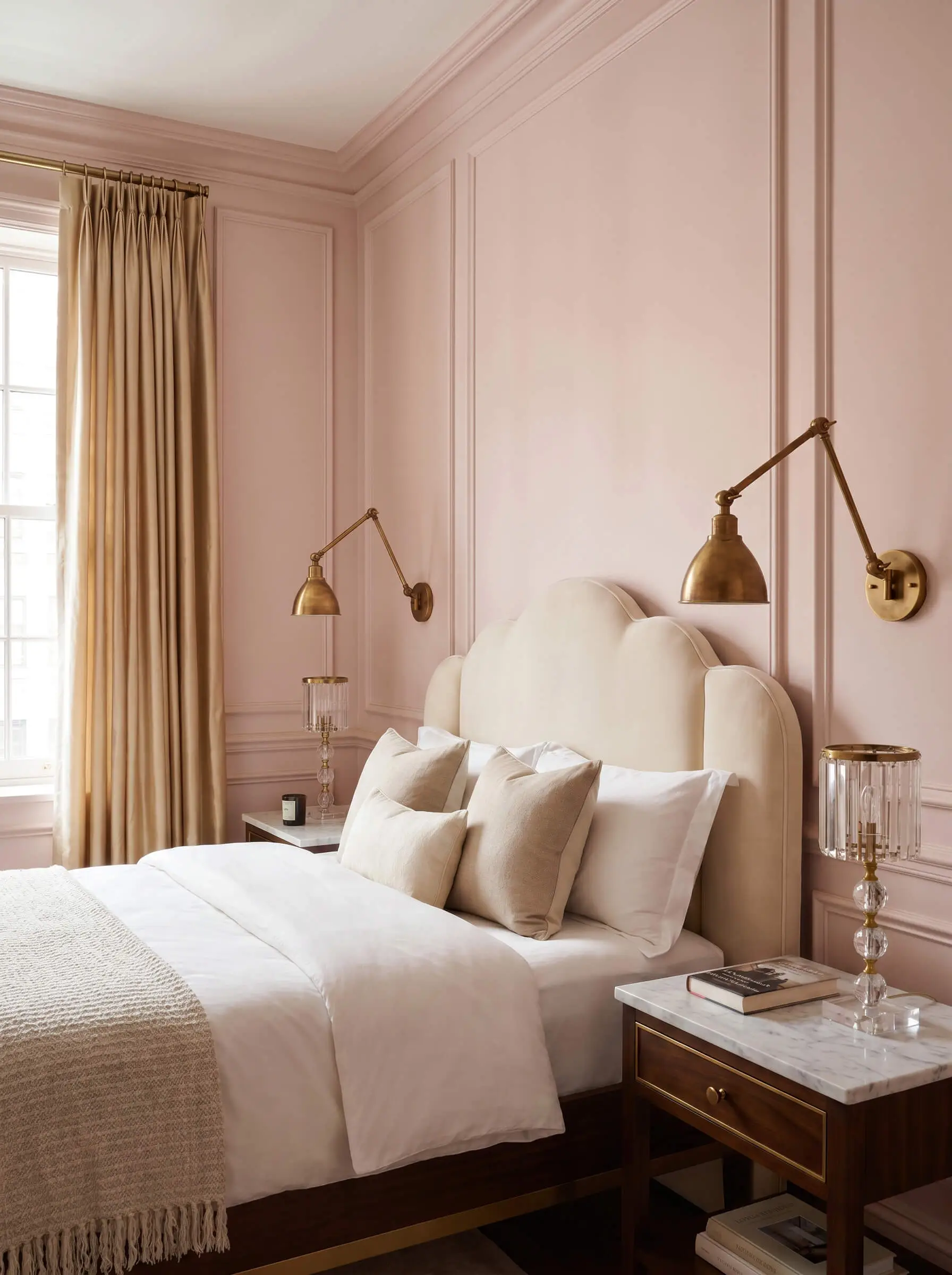 Solid unlacquered brass articulating sconces flank a custom scalloped headboard above marble-topped nightstands against soft pink architectural walls.