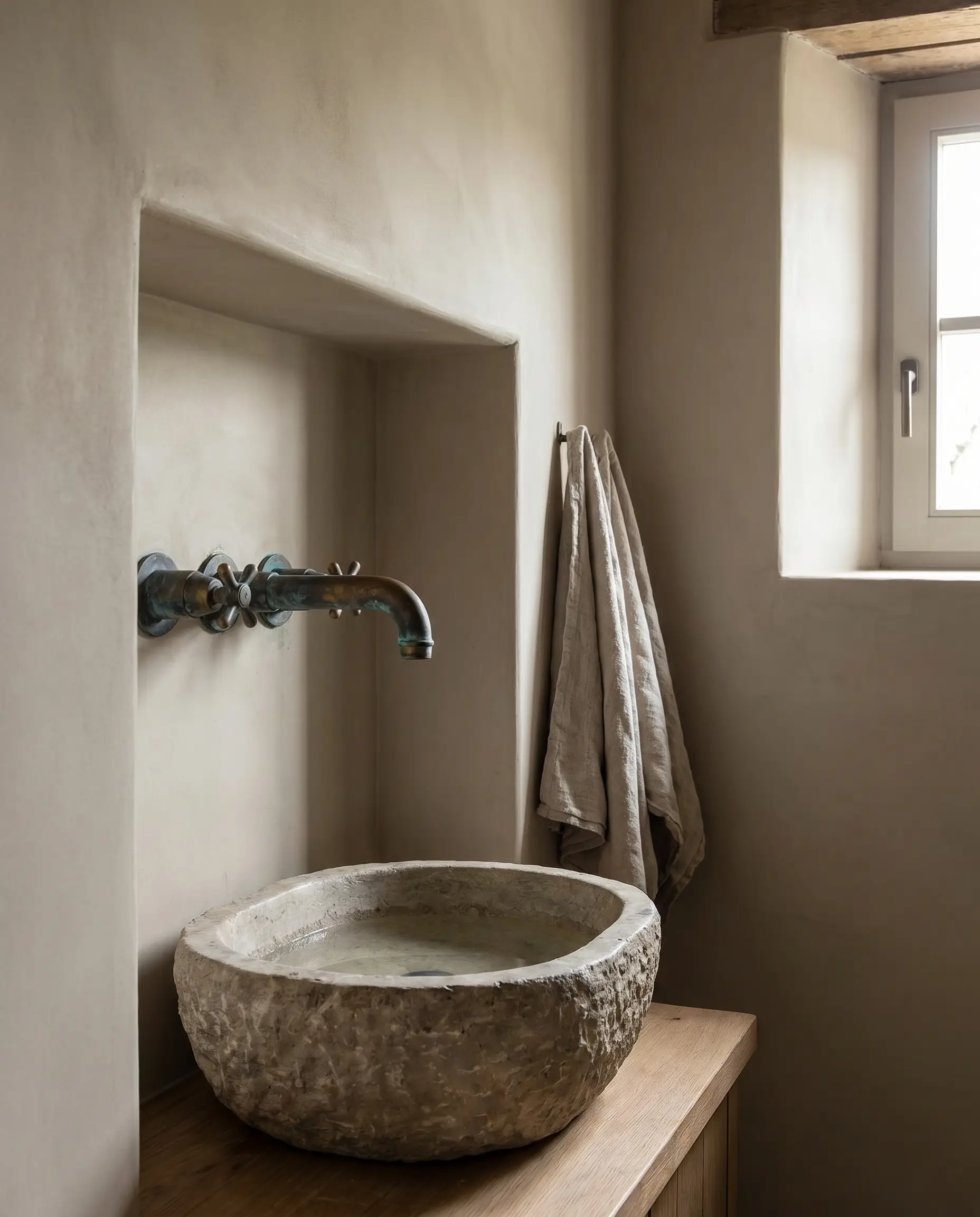 A solid unlacquered brass wall-mounted faucet with a rich living patina extends over a modest stone basin against smooth earthy greige bathroom walls.