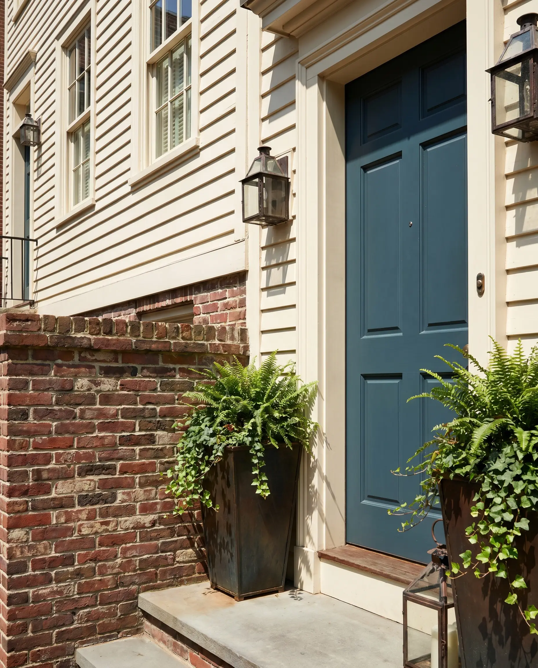 A solid front door painted in a deep gray-blue chromatic profile anchors a townhouse facade featuring creamy white siding, natural brick accents, and blackened steel planters.