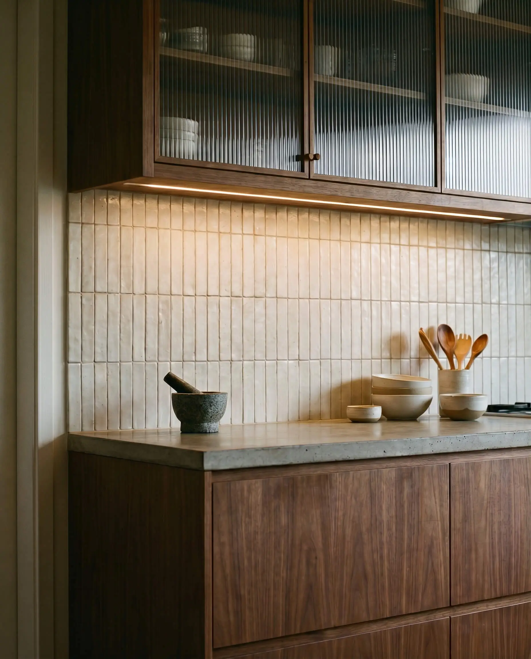 A matte handcrafted ceramic tile backsplash installed in a strict vertical grid sits between ribbed glass uppers and milled rift-sawn bases.