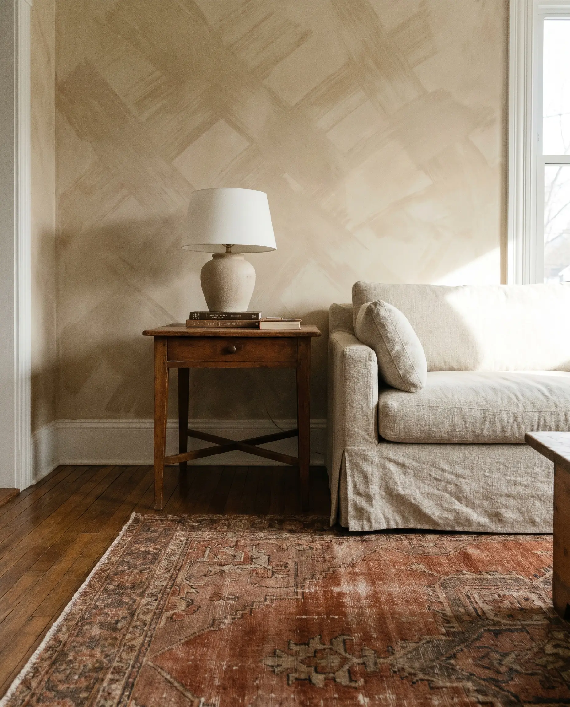 A faded vintage Oushak rug layered over hardwood floors counterbalances the sweeping cross-hatch strokes of sandstone limewash walls behind a wood side table.