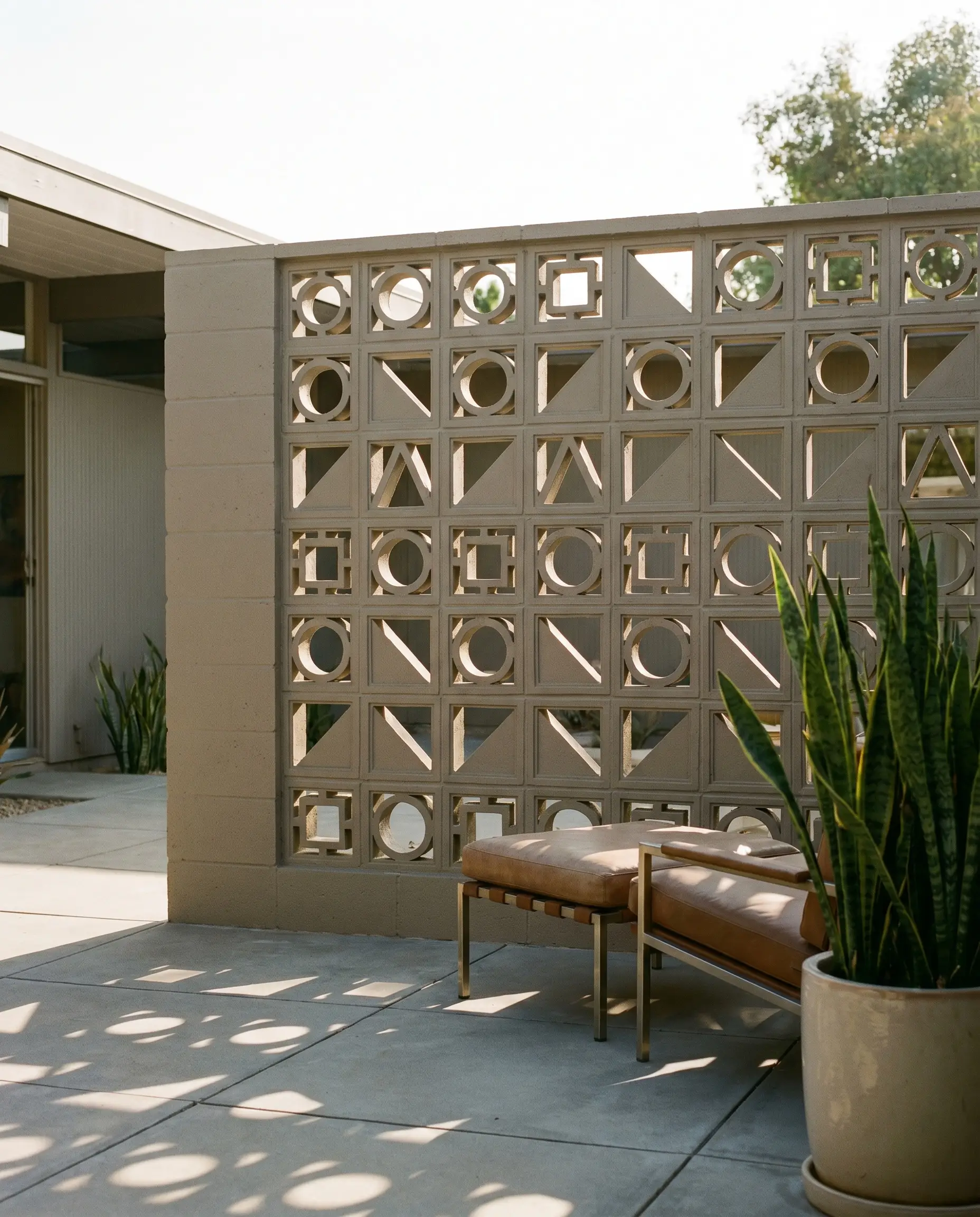 A geometric concrete breeze block privacy wall painted in a warm greige divides an outdoor courtyard featuring large-format paving stones and a modern lounge chair.