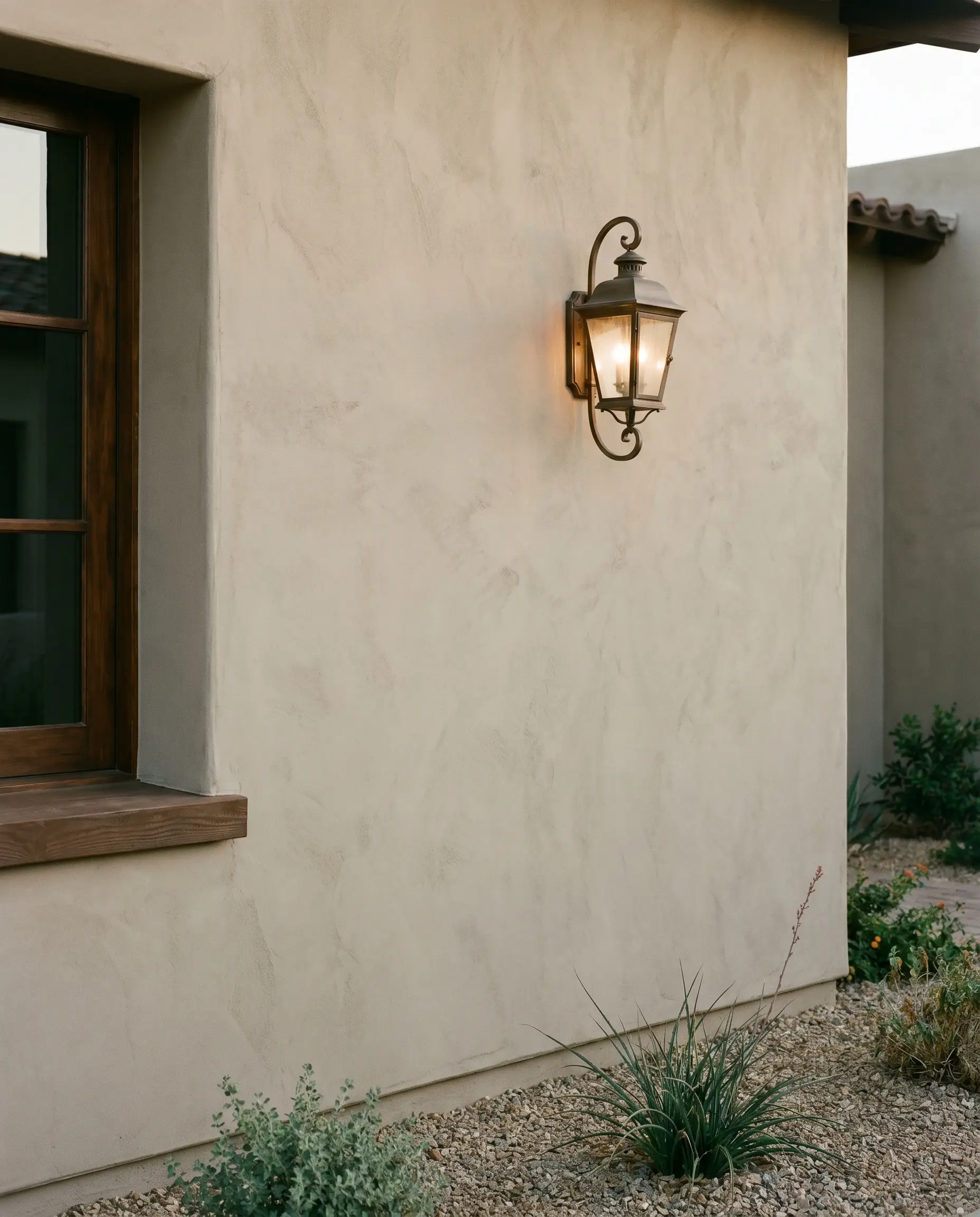 A flat-finish stucco wall coated in warm gray exterior paint pairs with an aged bronze lantern sconce and a dark wooden window frame.