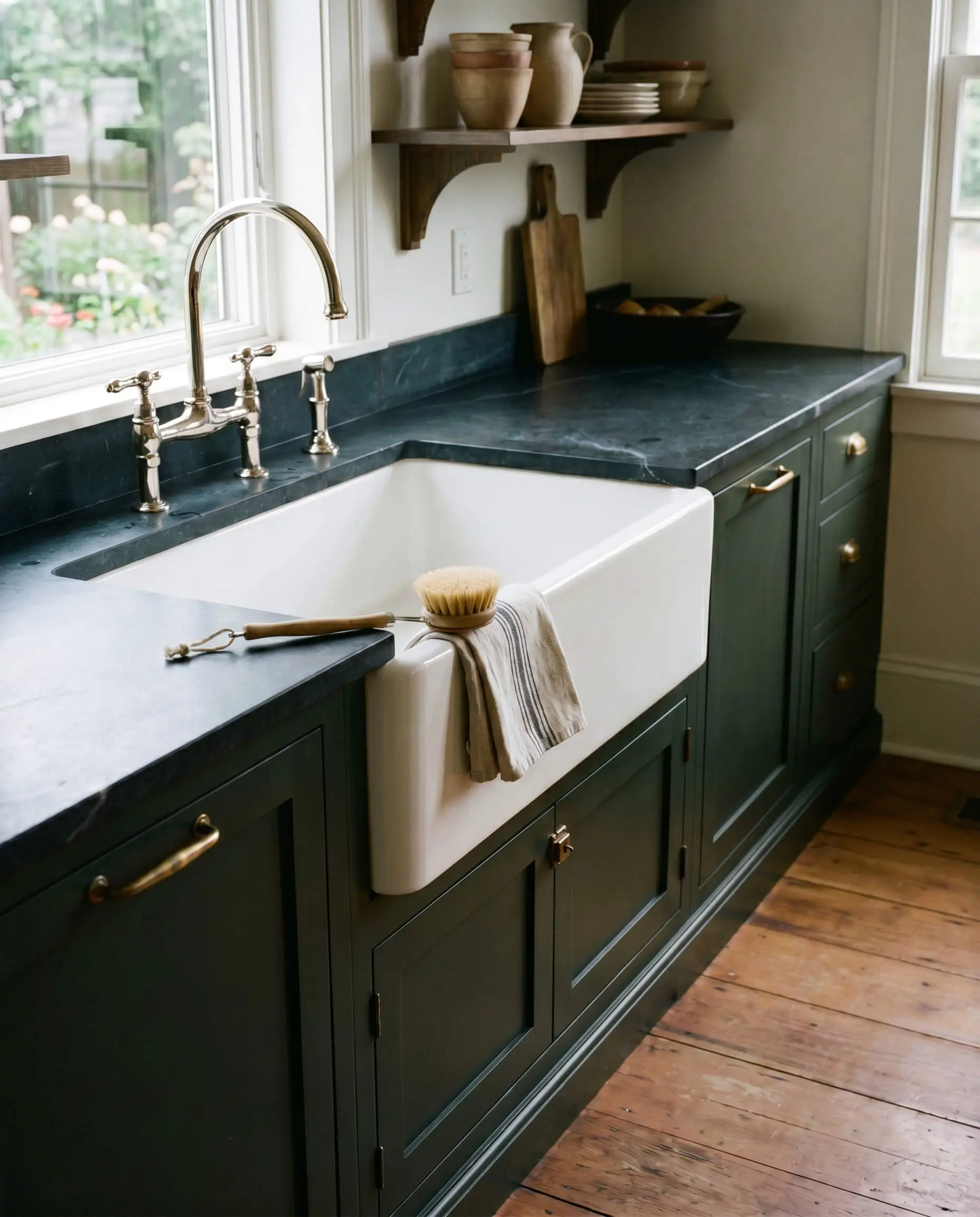 A massive white fireclay apron-front farmhouse sink breaks up Benjamin Moore Essex Green cabinetry, paired with undermounted soapstone countertops and a polished nickel faucet.
