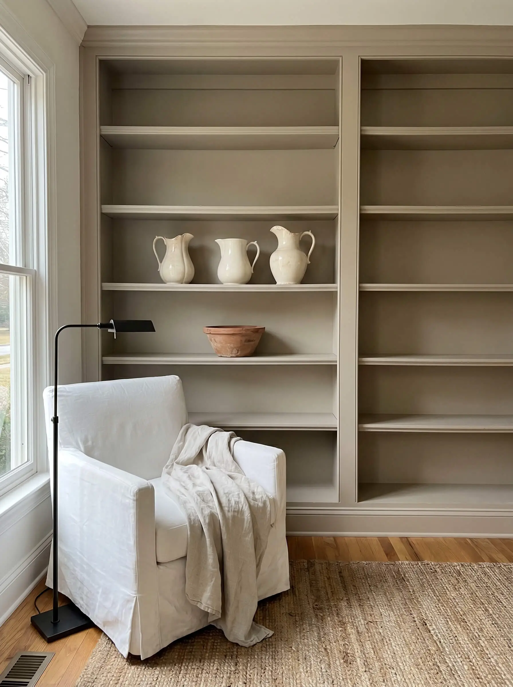 Three antique ironstone pitchers and a raw terracotta bowl rest on muddy neutral bookshelves near a modern slipcovered armchair and an iron reading lamp.