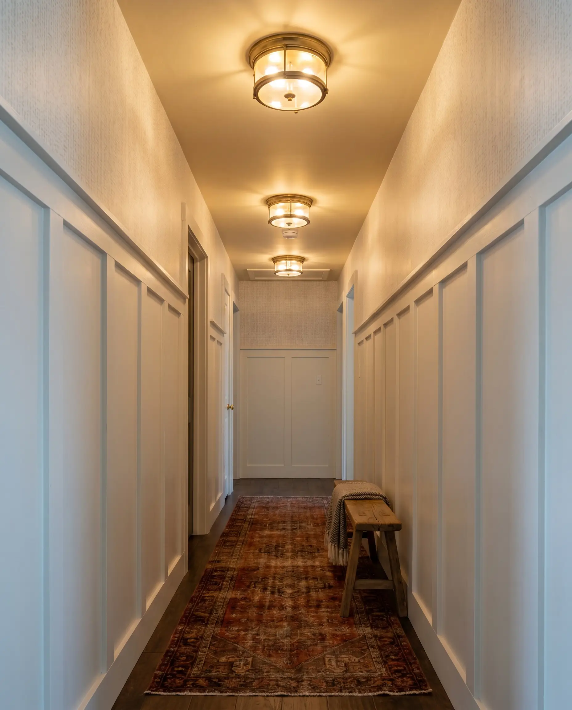 A wooden entryway bench and a vintage runner rug line a windowless hallway featuring board-and-batten wainscoting and a buttery off-white ceiling.