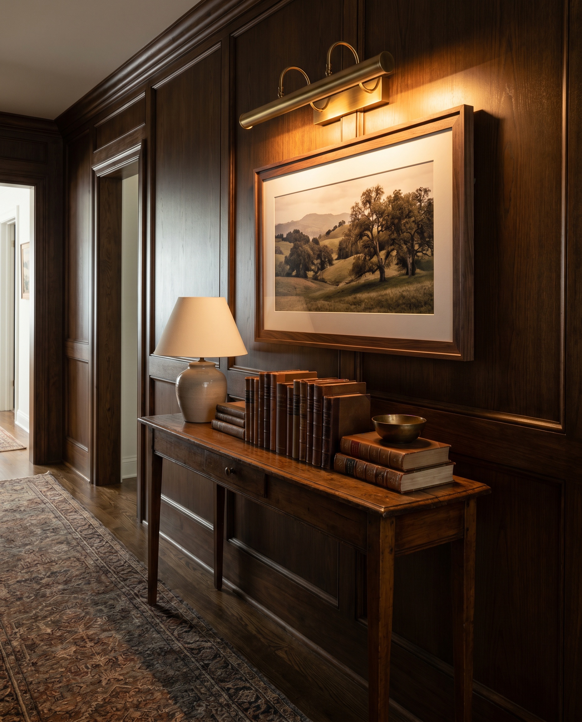An unlacquered brass picture light casts a warm glow over a landscape poster in a dark wood frame above a heritage hallway console table.