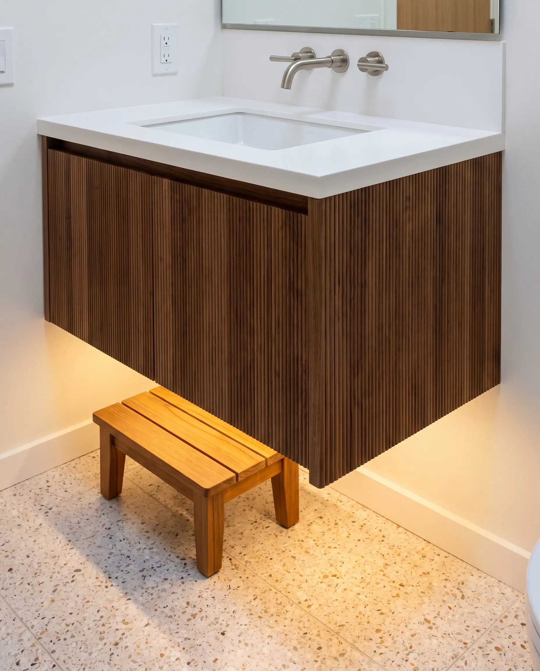 A cantilevered ribbed walnut floating vanity with a white integrated sink sits above continuous terrazzo floor tiles and a minimal teak step stool.