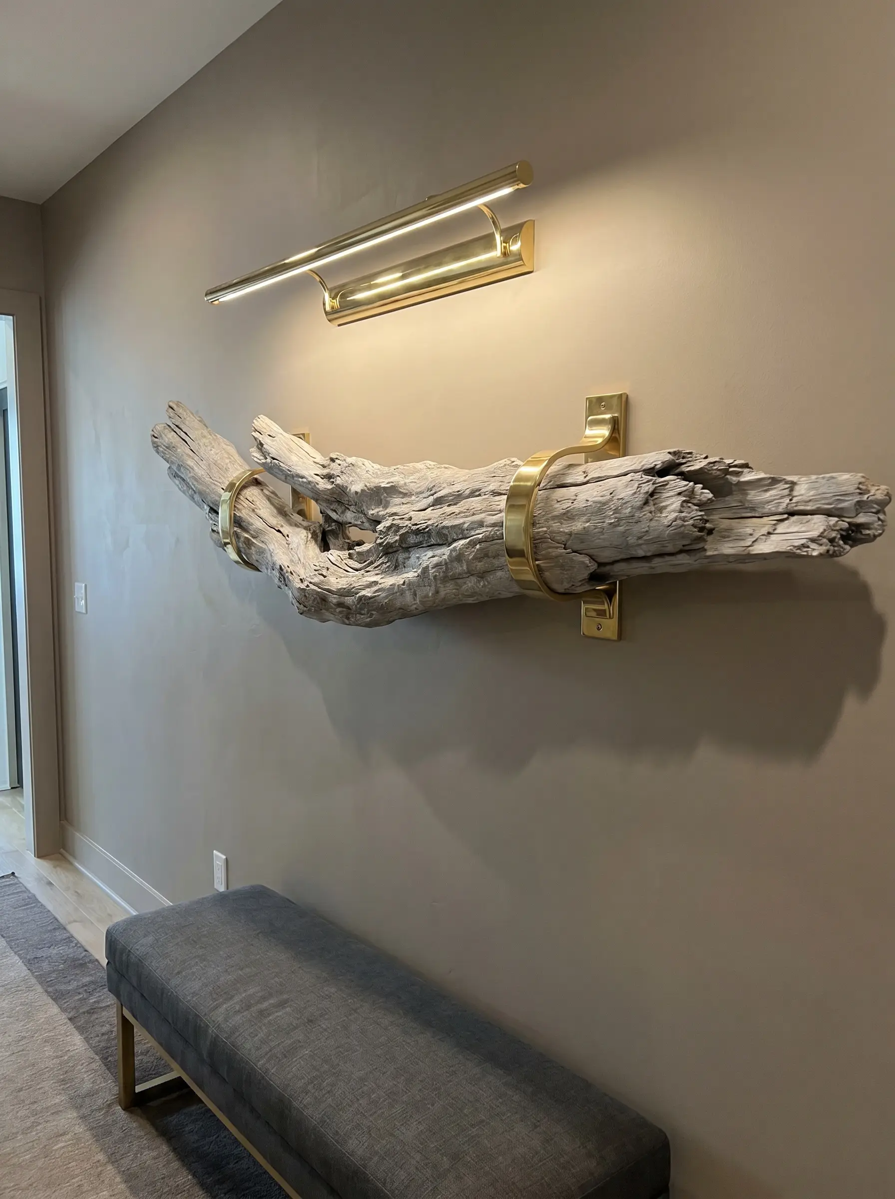 Highly weathered silvery gray driftwood wall art is secured by solid brass brackets and illuminated by an overhead polished brass picture light.
