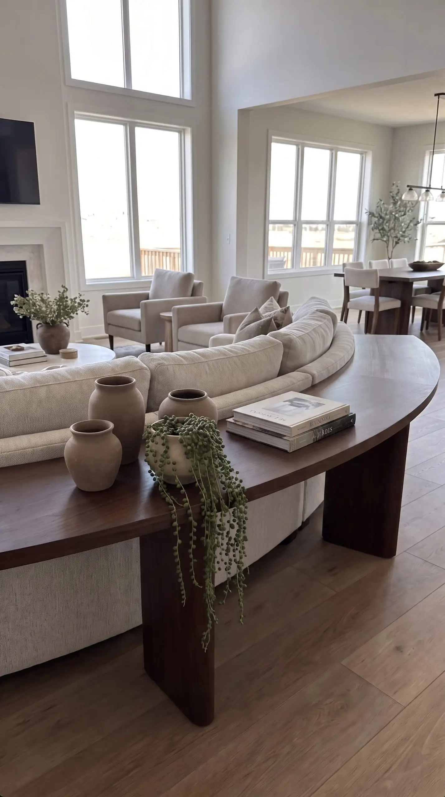 A sweeping curved console table mathematically hugs the backrest of a floating serpentine sofa, styled with matte ceramic vessels, heavy art books, and trailing plants.