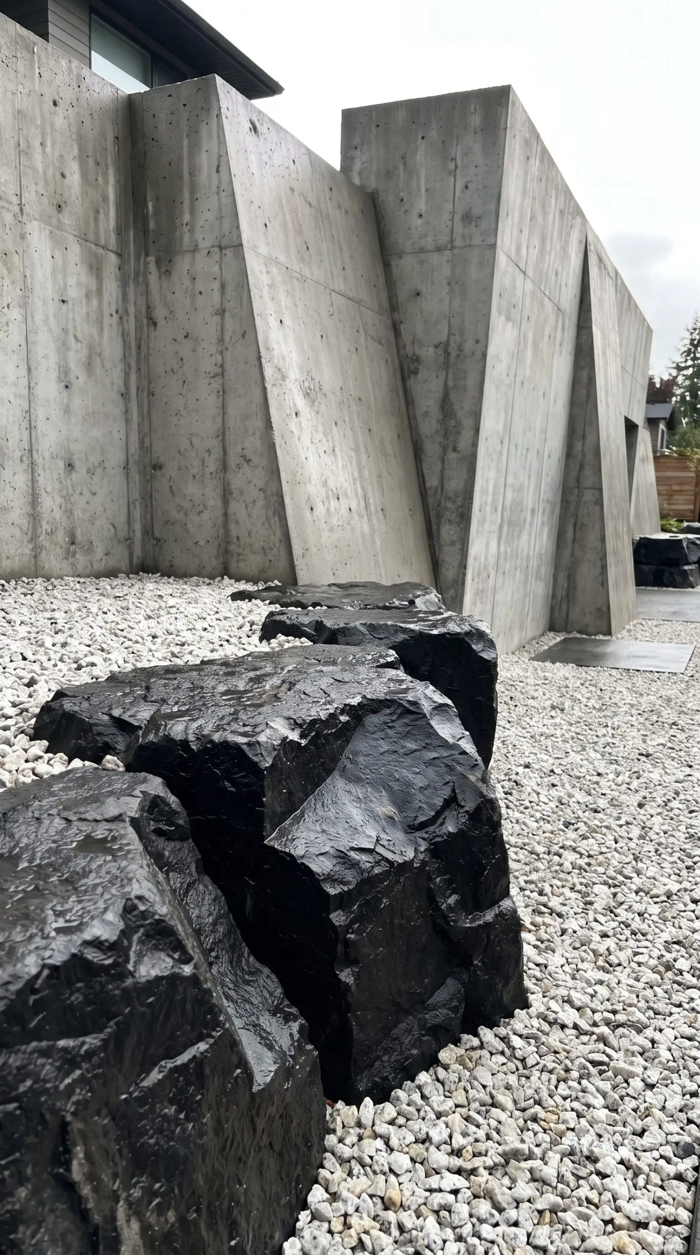 Deep charcoal-toned raw basalt boulders contrast sharply against a bright pale crushed granite base in front of a sleek concrete retaining wall.