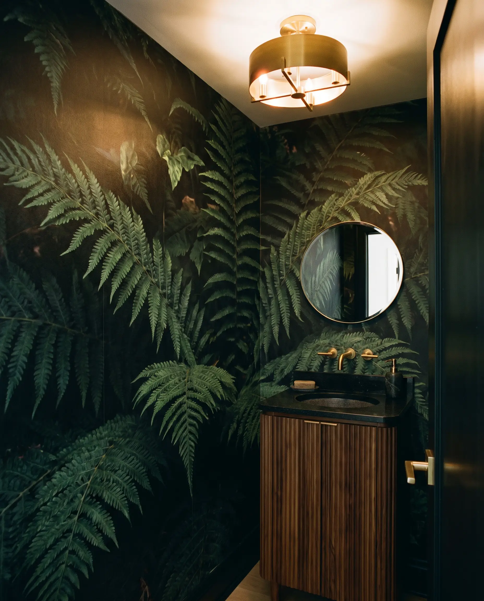 A narrow freestanding ribbed wood vanity with a black granite top sits against an immersive dark botanical panoramic wallpaper mural featuring moody green ferns.