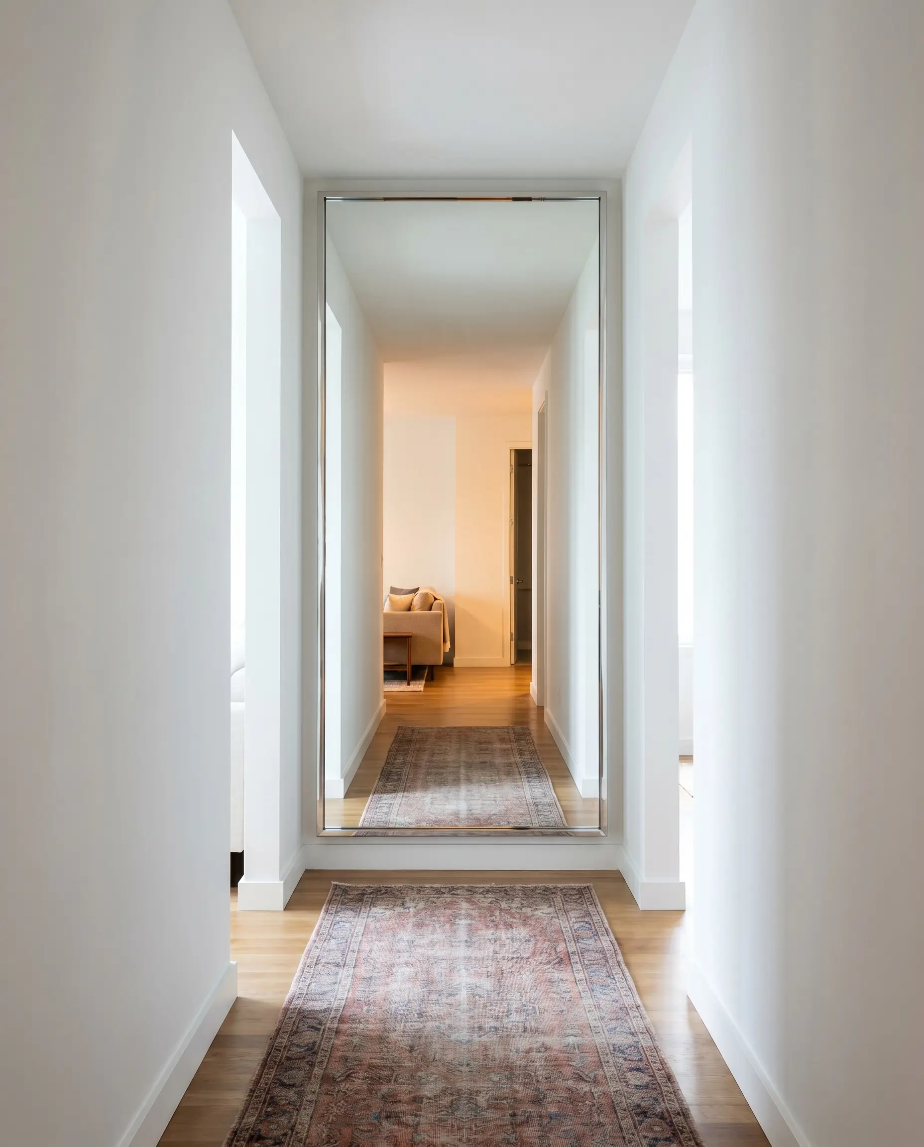 An unframed polished-edge floor-to-ceiling mirror anchors the terminus wall as narrow hallway wall decor, reflecting a vintage runner rug.