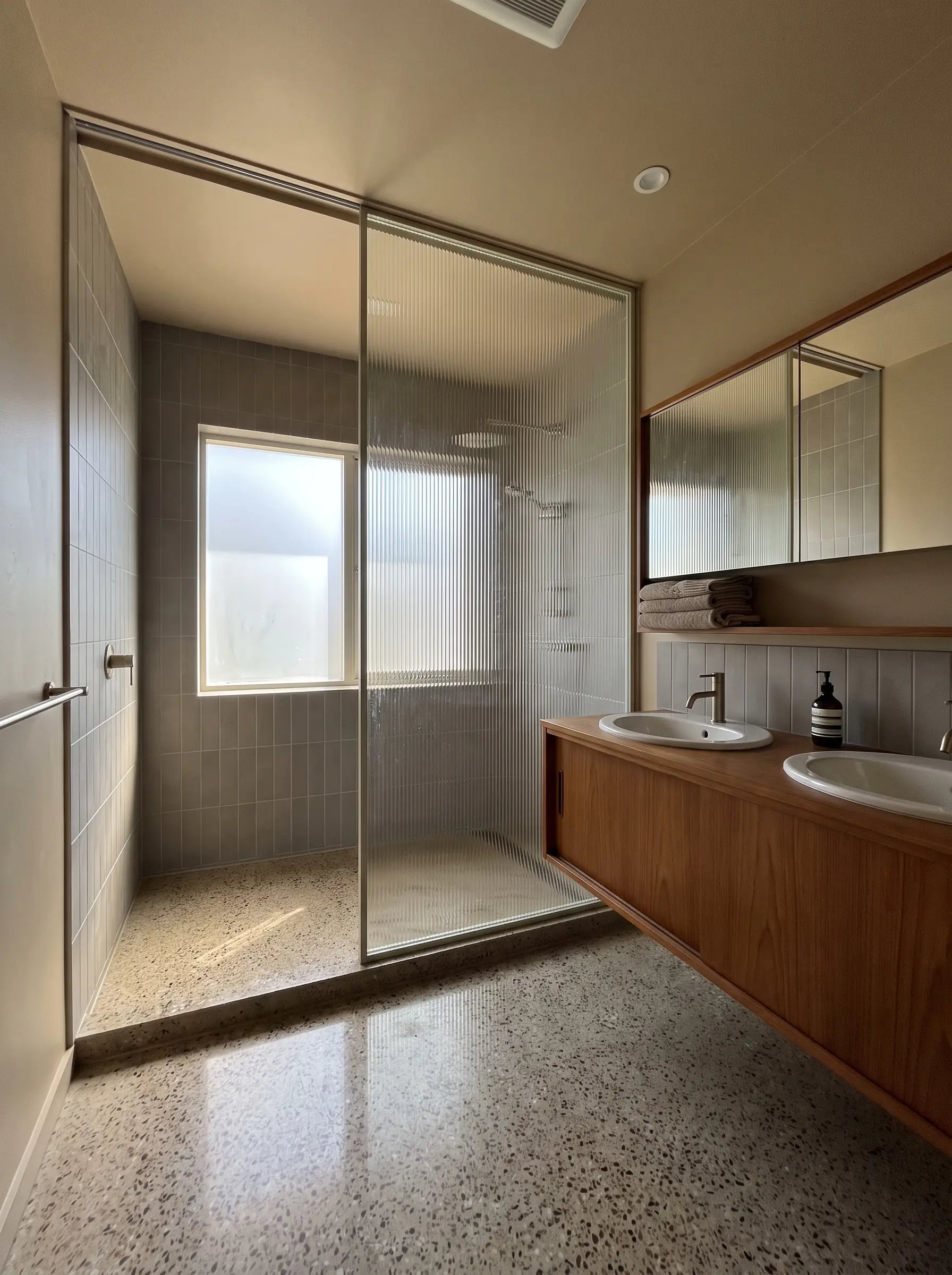 A thick fluted architectural glass panel is recessed into the ceiling and floor via a seamless channel beside a warm wood bathroom vanity.