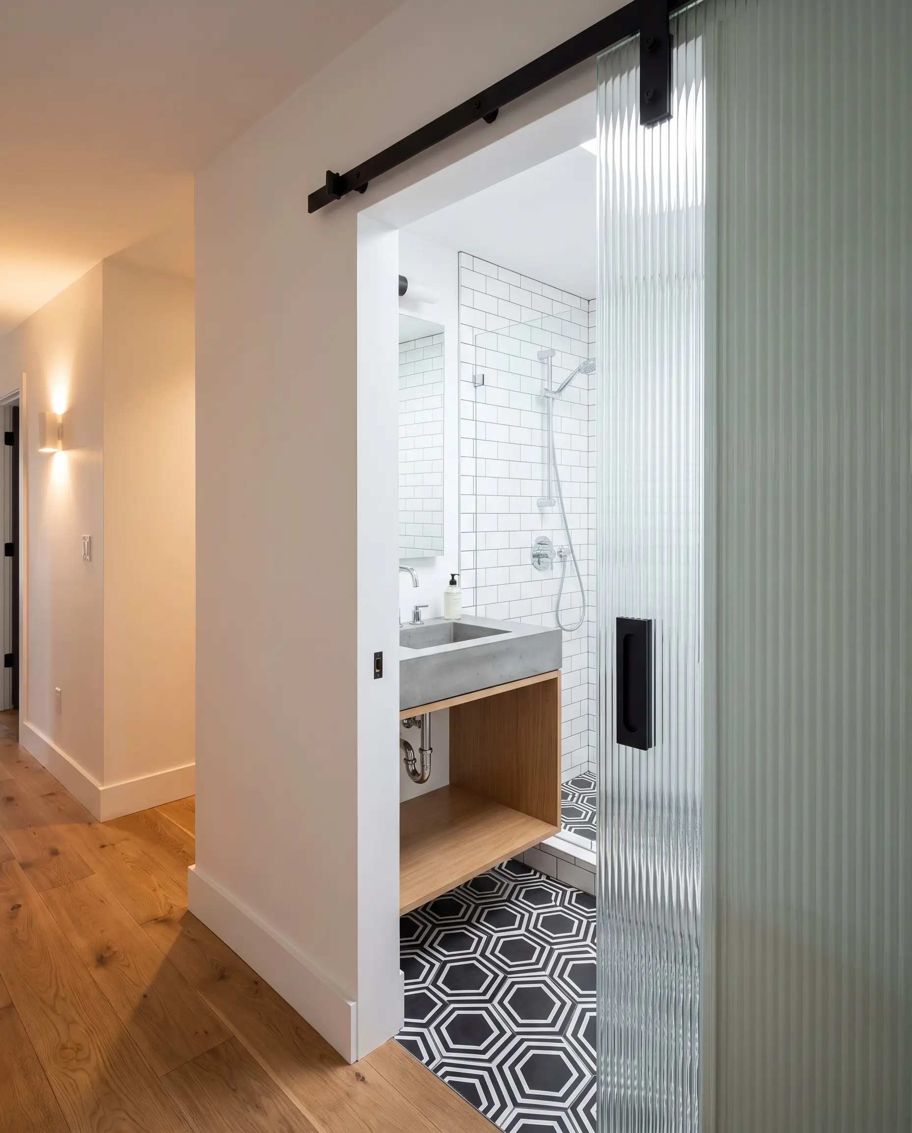 A fluted glass barn door on matte black track hardware slides open to reveal a narrow-depth floating vanity and geometric black and white hex tiles.