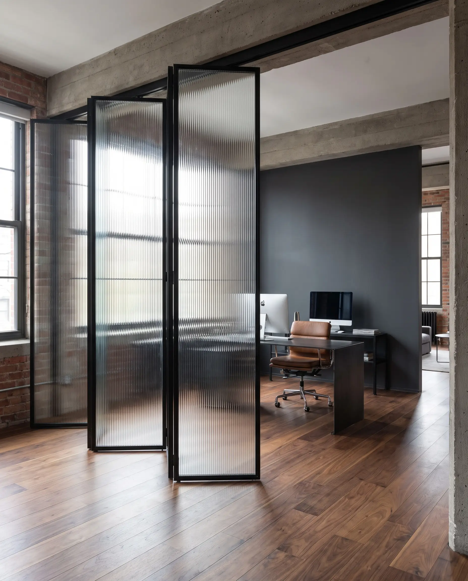 Heavyweight ribbed glass panels set in blackened steel frames stand on wide-plank walnut flooring, obscuring a sleek desk against a Farrow and Ball Railings accent wall.