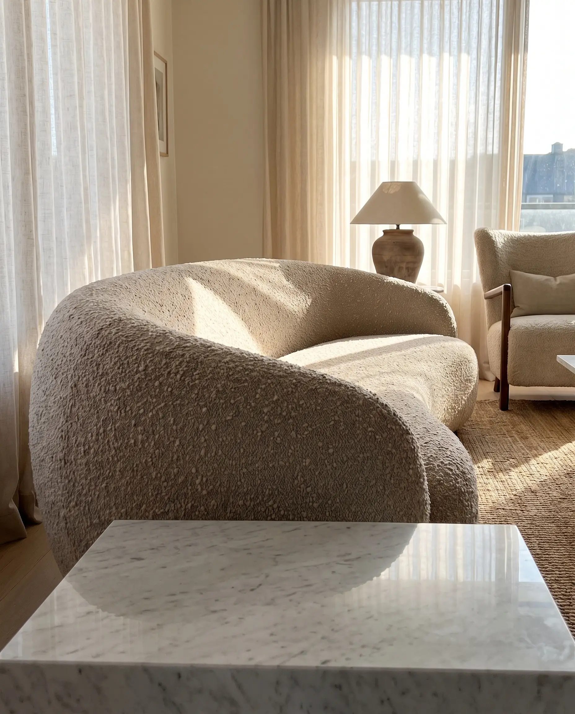 A warm oat heavyweight wool bouclé curved sofa design contrasts sharply against a smooth, cold polished marble coffee table in an approachable luxury living space.