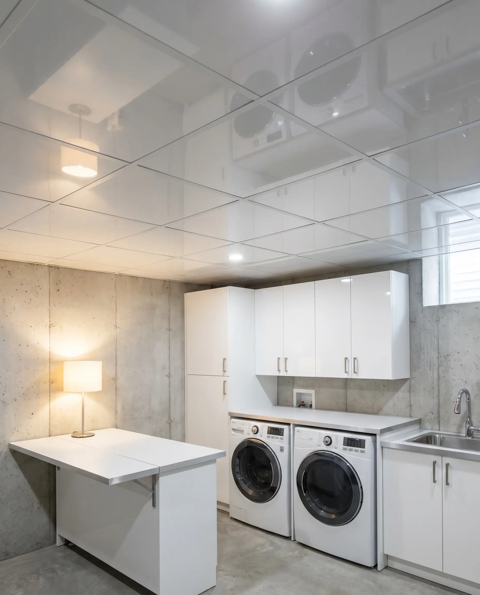 Rigid high-gloss white PVC lay-in panels form a waterproof suspended grid system above a modern basement laundry zone featuring sleek cabinetry and a folding counter.