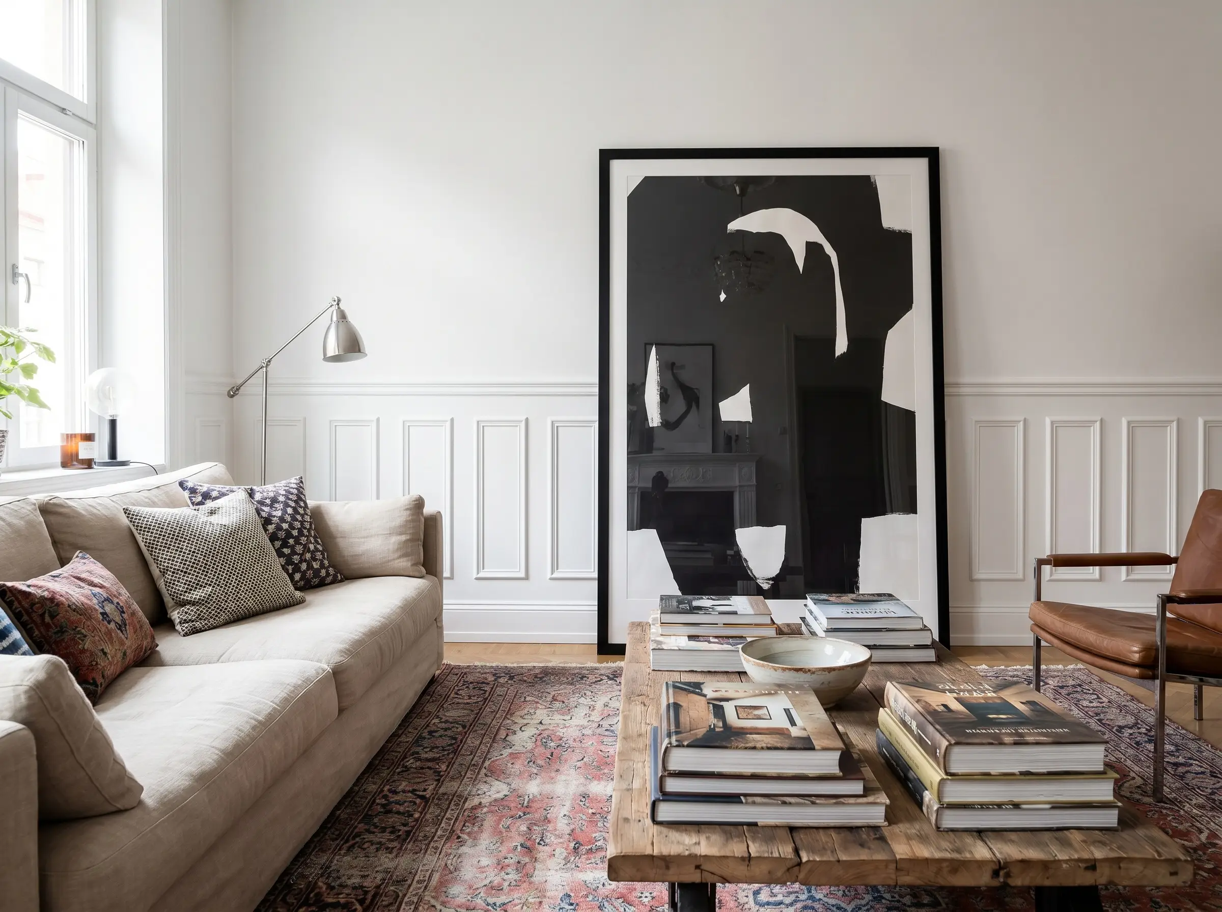 Interior decorating using posters showcases an oversized contemporary art print in a black frame intentionally overlapping traditional white wainscoting behind a neutral sofa.