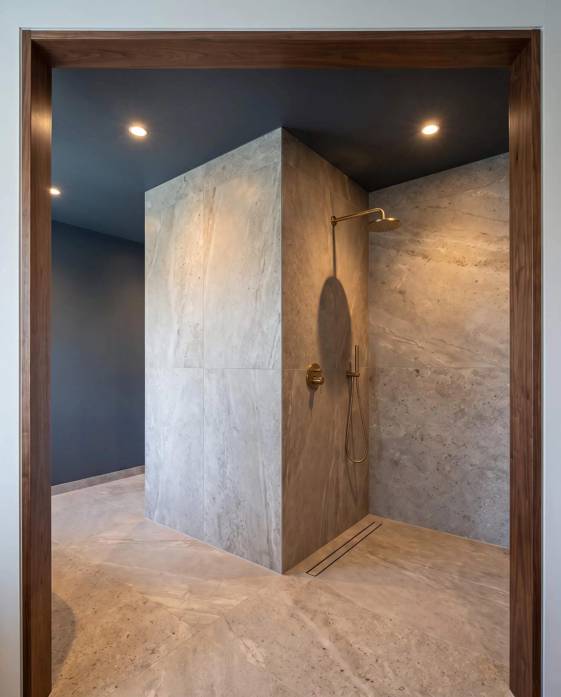A structural wall clad in large-format porcelain slabs forms an L-shaped corner baffle beneath a Farrow and Ball Hague Blue painted ceiling and brass plumbing fixtures.