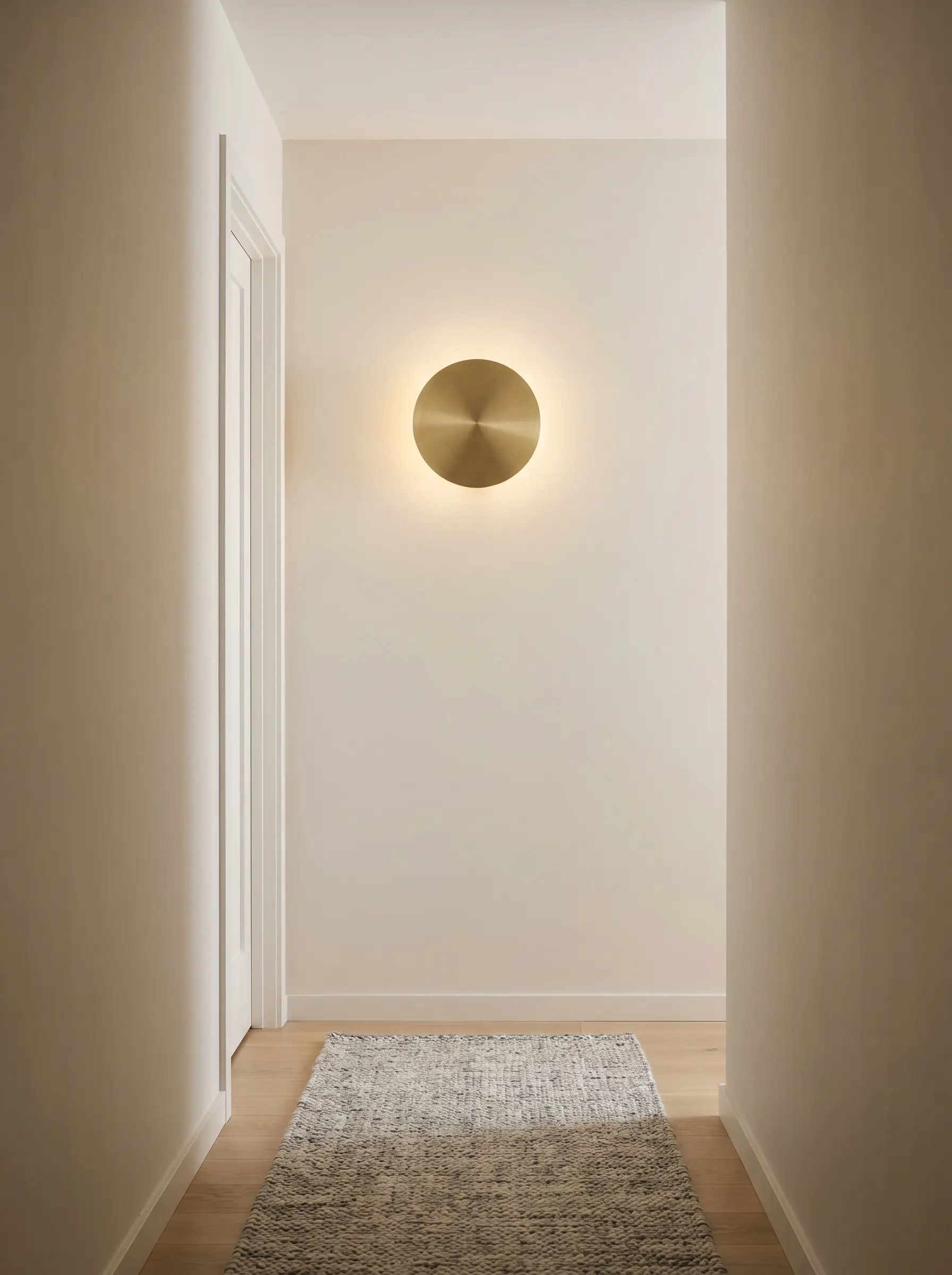 A sleek brass disc sconce sits flush against a smooth neutral wall, providing an ADA-compliant low-profile fixture next to a white door frame.