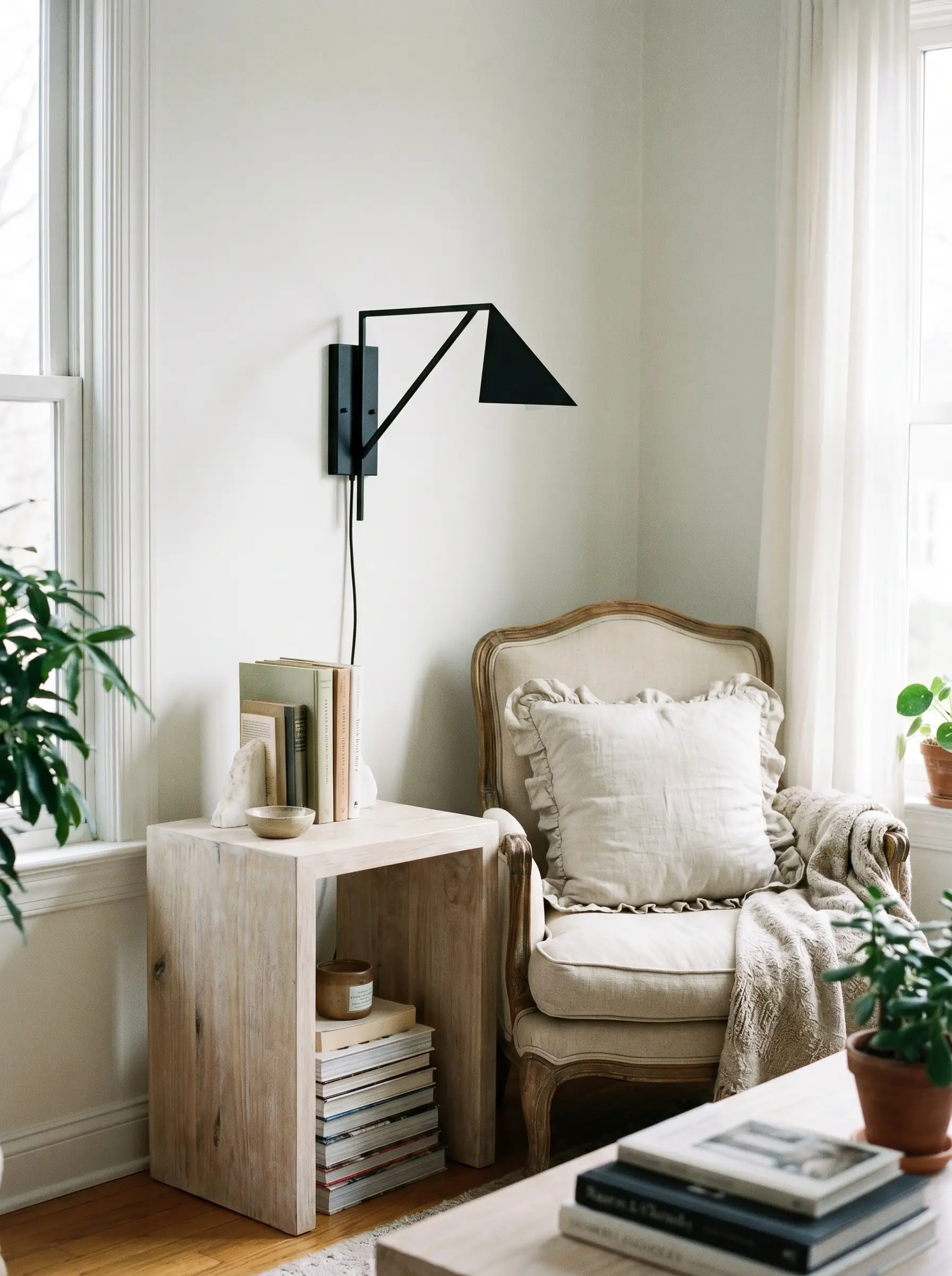 A geometric matte black iron wall sconce illuminates a bleached wood side table and an armchair styled with a ruffled linen pillow.
