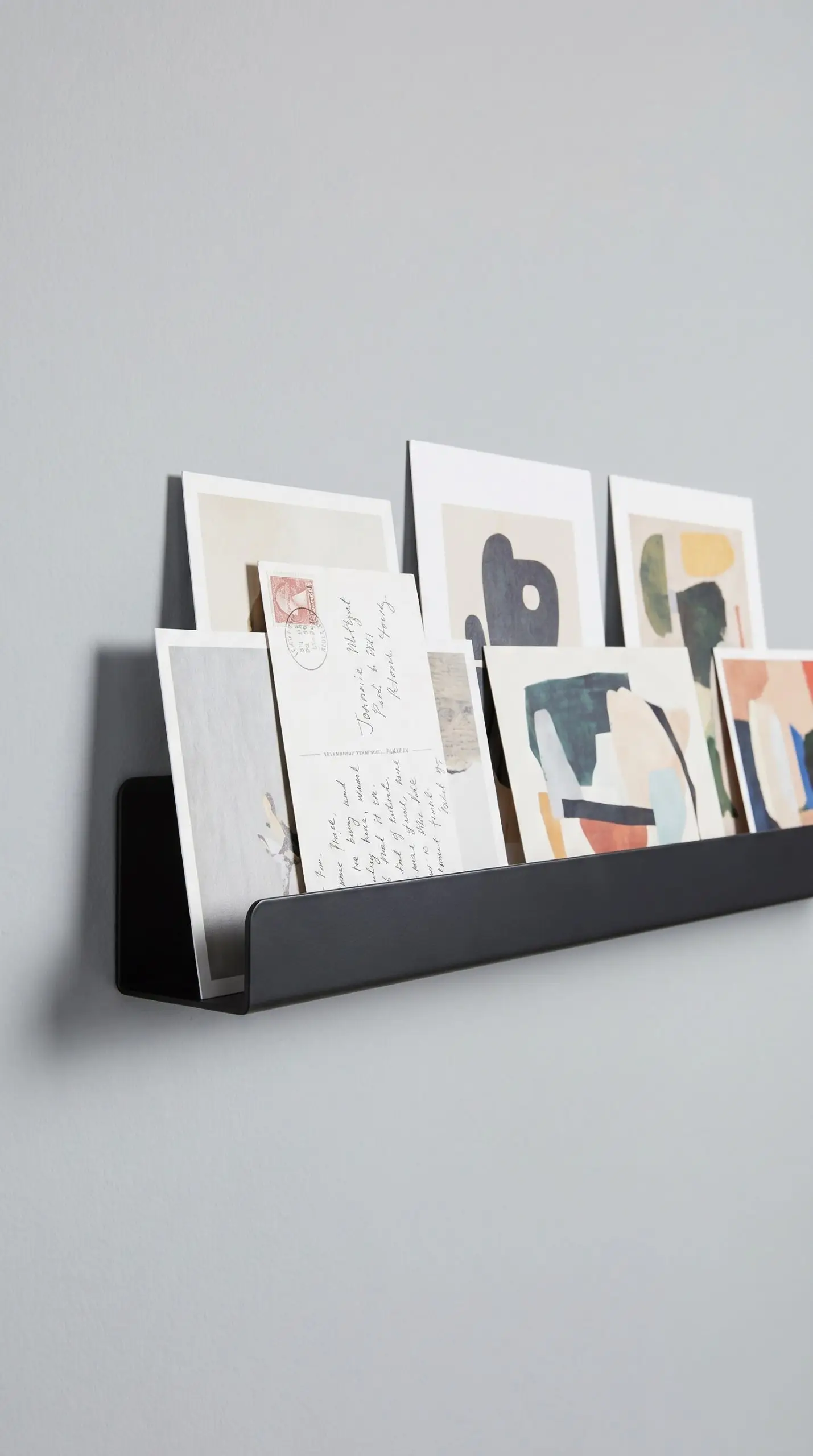 A two-inch deep powder-coated black steel picture ledge holds overlapping unframed prints against a soft gray wall as streamlined narrow hallway wall decor.
