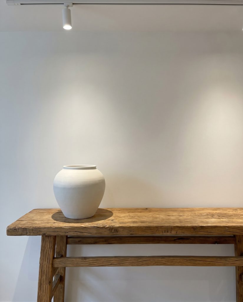 A single oversized, unglazed white ceramic stoneware vessel rests off-center on an antique, reclaimed pine wood console table in a wabi-sabi entryway.
