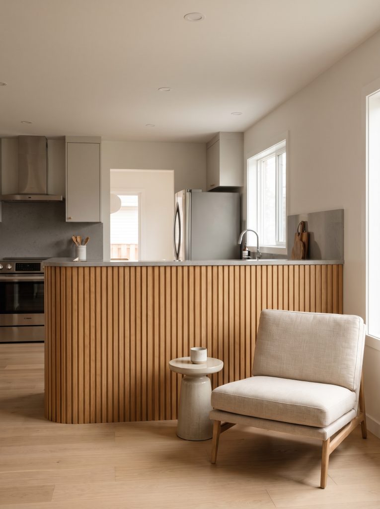 Vertical rift-sawn white oak slats wrap the living room facing side of a kitchen partition, contrasting with stainless steel appliances and quartz countertops.