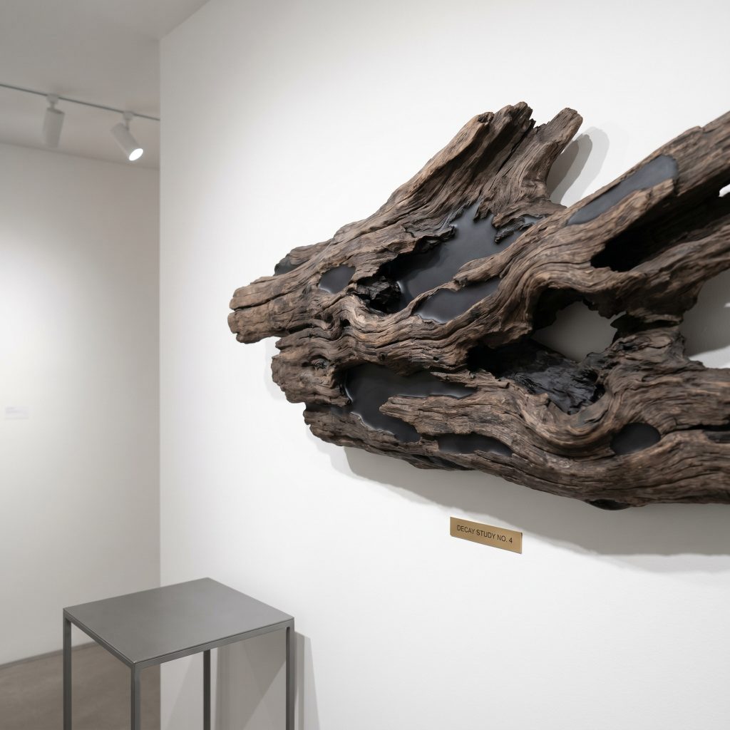 A sculptural driftwood piece featuring natural cracks filled with matte charcoal epoxy resin hangs on a pristine white gallery wall above a contemporary side table.