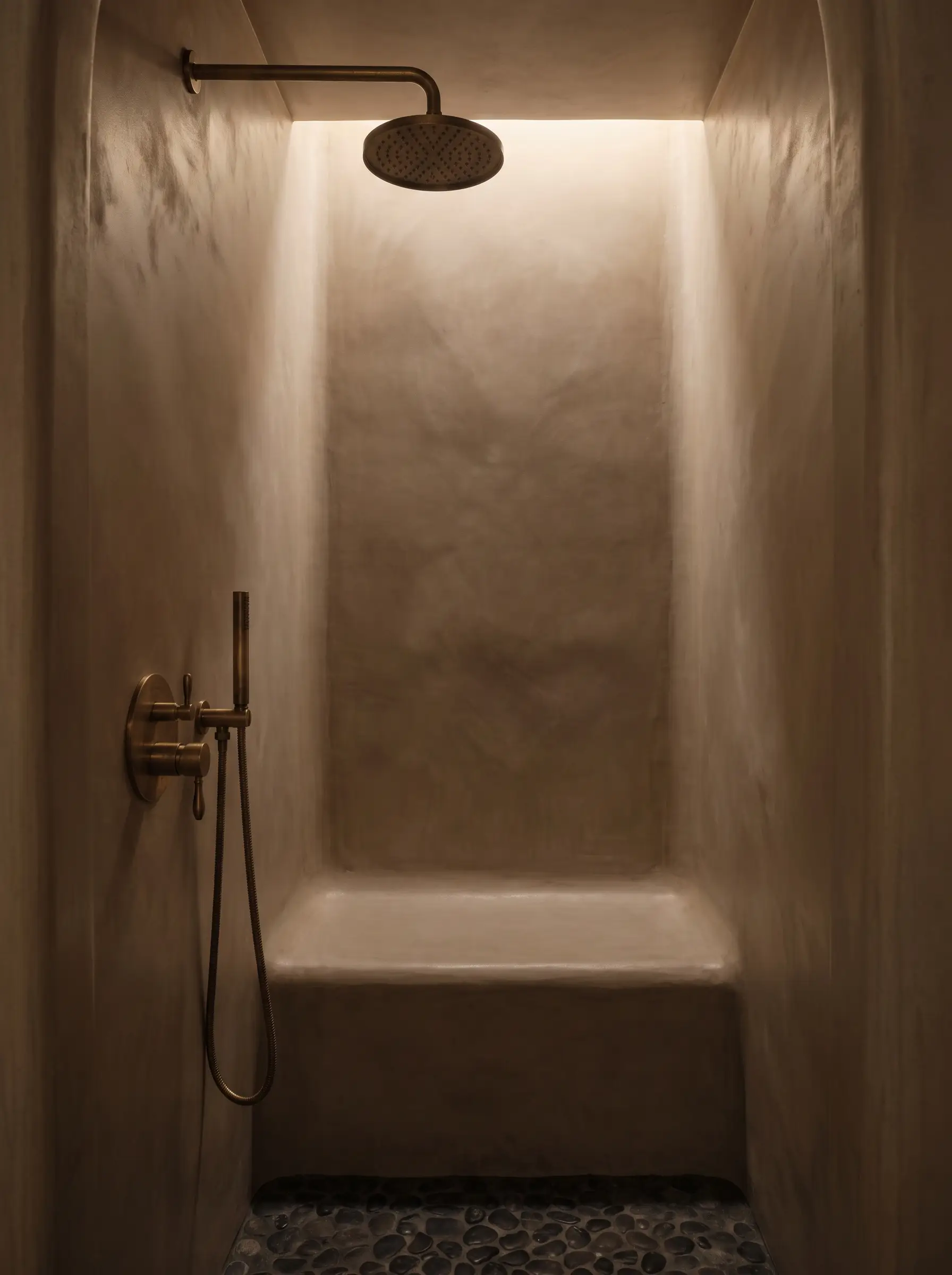 A seamless shower alcove coated in warm mushroom Tadelakt plaster features a built-in bench and unlacquered brass rainfall shower fixtures.