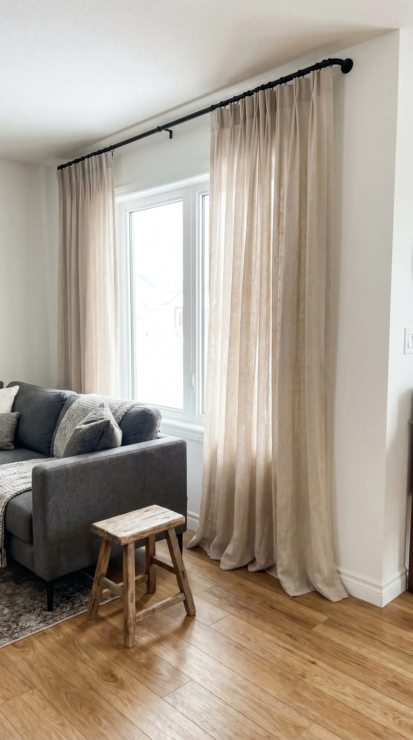 Flowing pinch-pleated sheer linen drapes hang from a matte black iron French return rod beside a sharp modern sofa and a vintage milking stool.