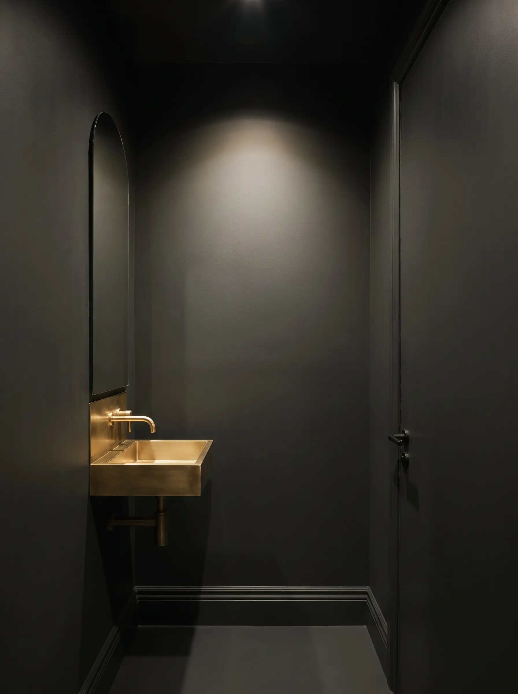A small bathroom design color-drenched in Sherwin-Williams Greenblack features a minimalist unlacquered brass wall-mounted sink beneath a tall slender arched mirror.