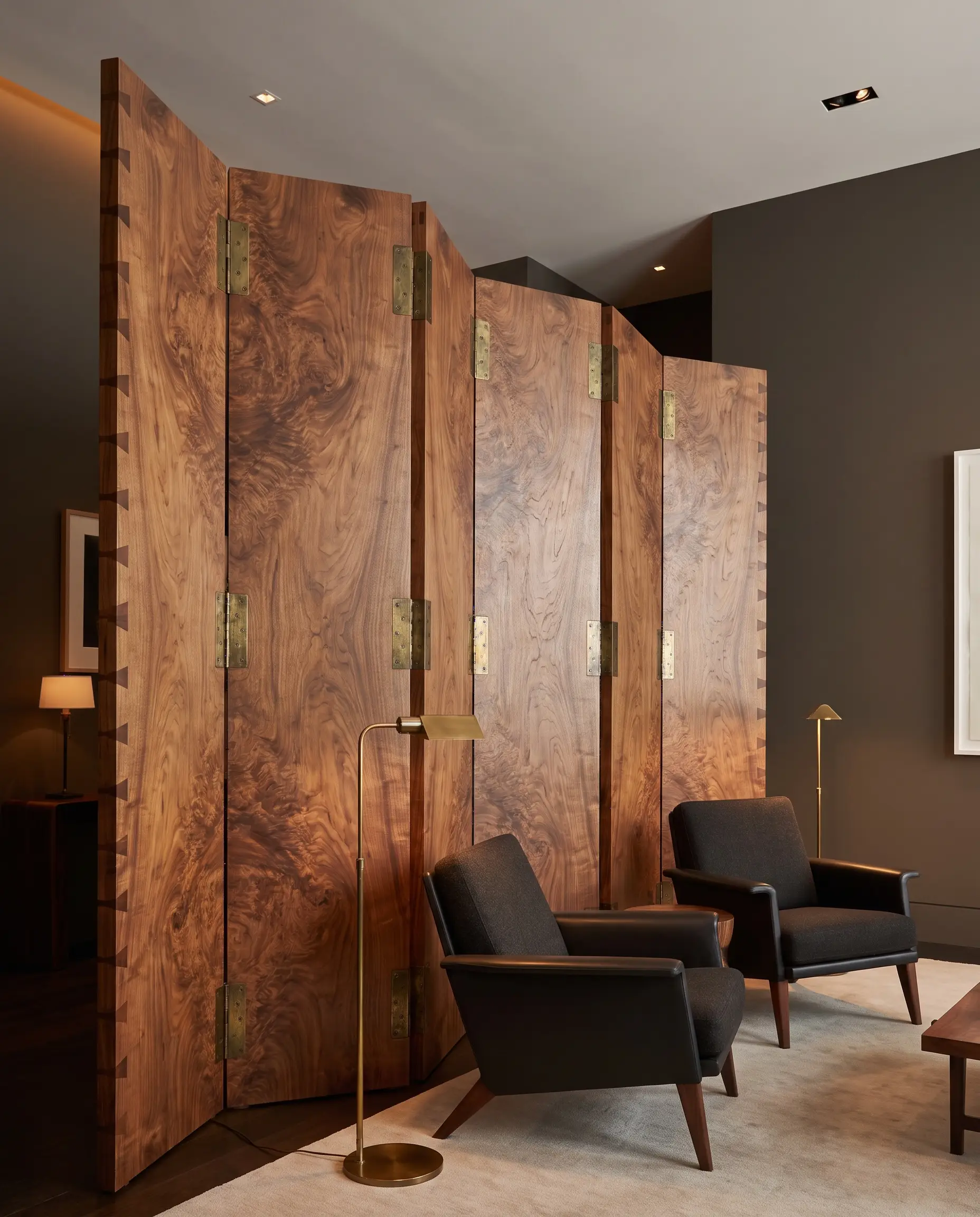 A massive solid burl wood paravent with exposed dovetail joinery and heavy brass hinges grounds a seating area featuring low-profile contemporary lounge chairs and Urbane Bronze walls.