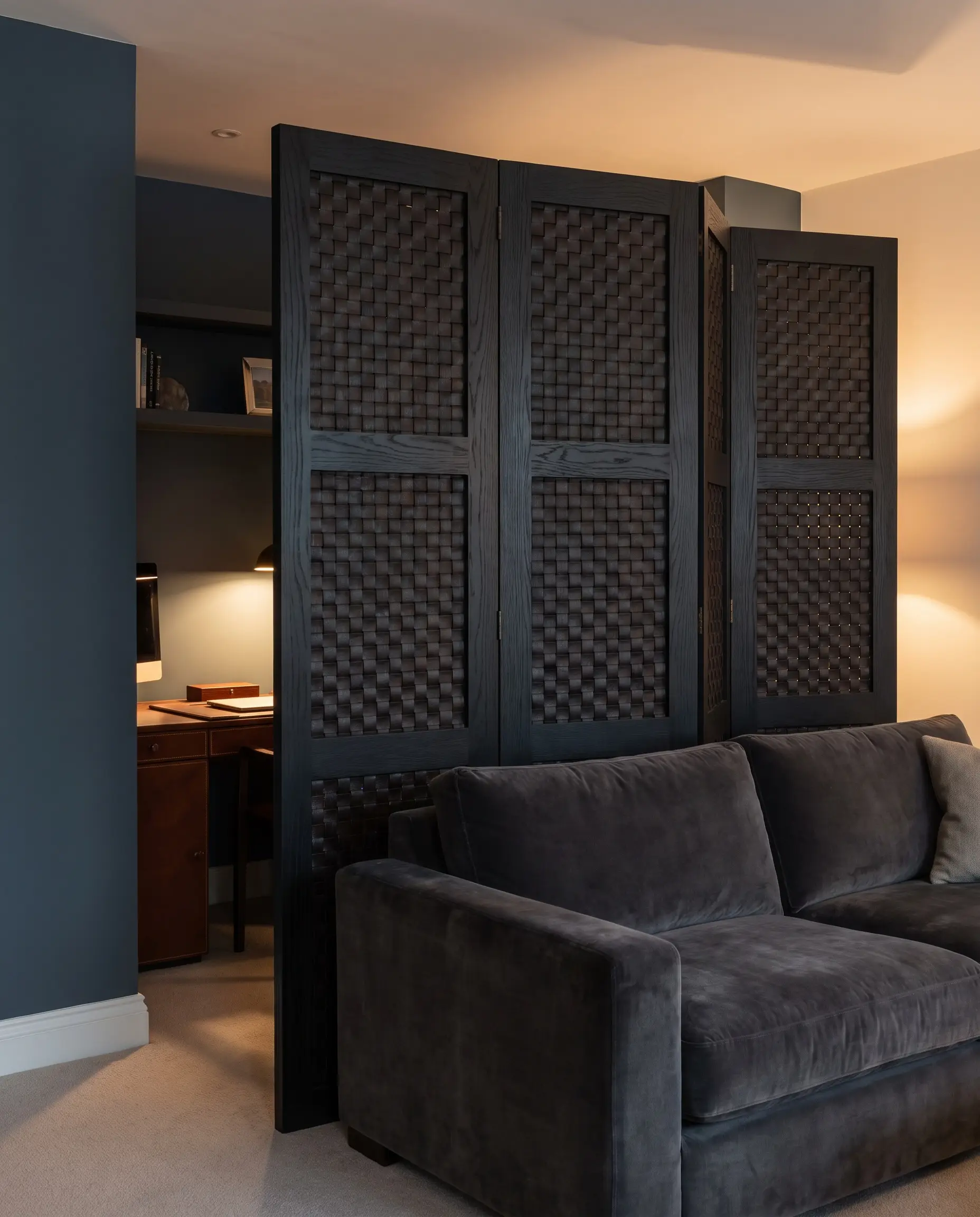 A six-foot tall ebonized oak and woven leather room divider completely obscures a home office desk from a luxurious living room sofa, backed by Hague Blue walls.