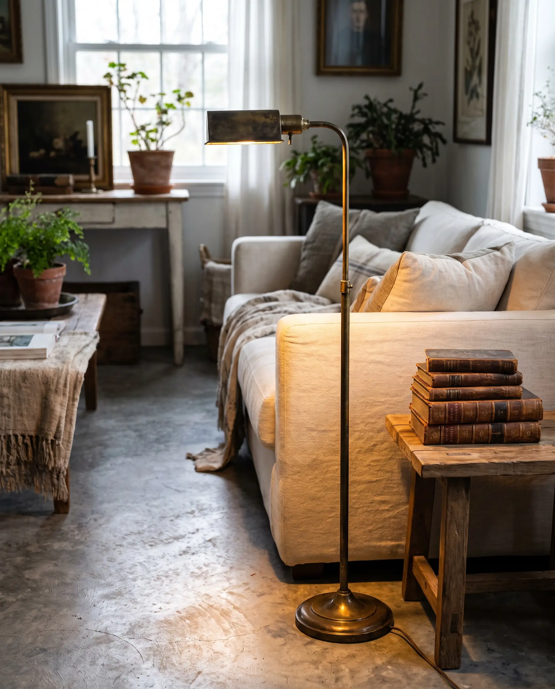 An unlacquered brass floor lamp casts a warm glow over a cream sofa and a stack of books in a cozy living room.