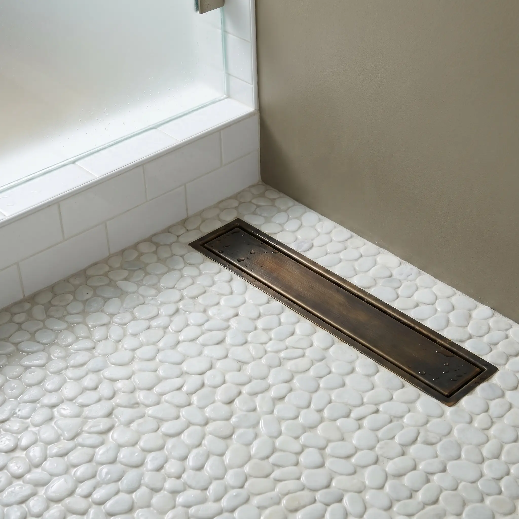 An unlacquered brass linear drain with a rich natural dark patina is set flawlessly into a pristine white pebble mosaic shower floor.