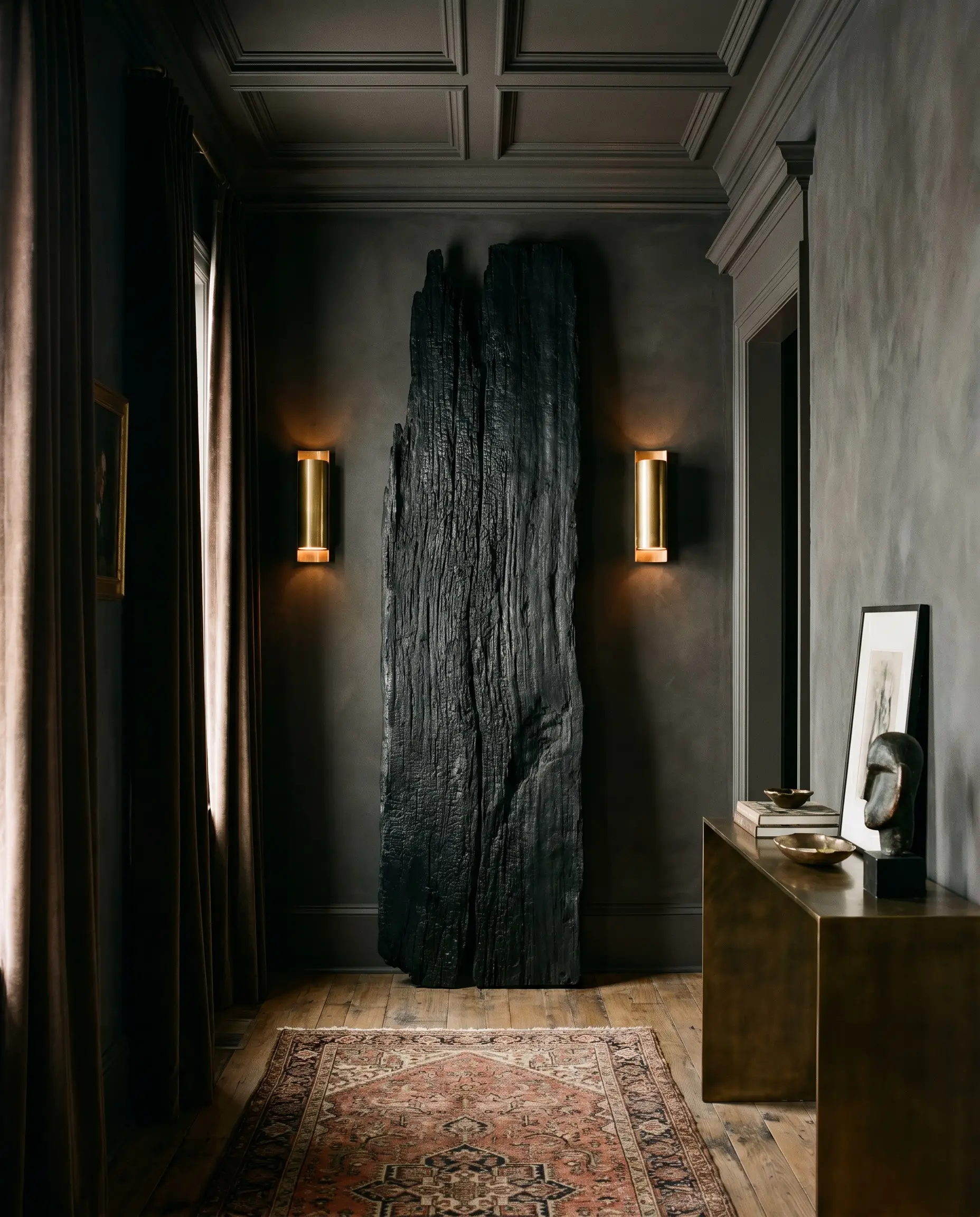A tall ebonized wood finishes driftwood slab anchors a narrow entryway flanked by unlacquered brass cylinder wall sconces.