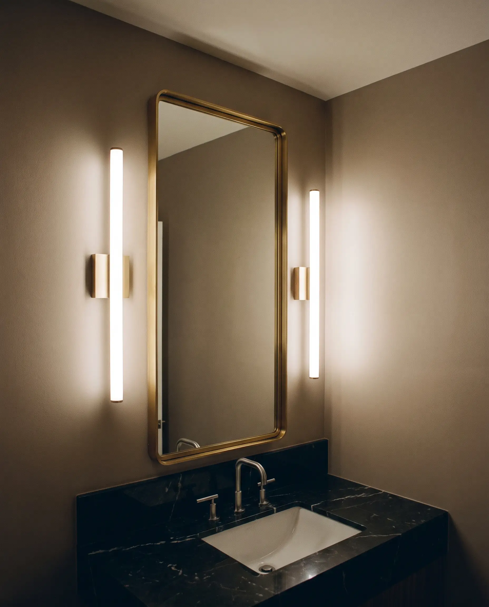 Slim frosted acrylic vertical tube sconces flank a tall brass-framed mirror above a sleek black marble vanity top and undermount sink against matte taupe walls.