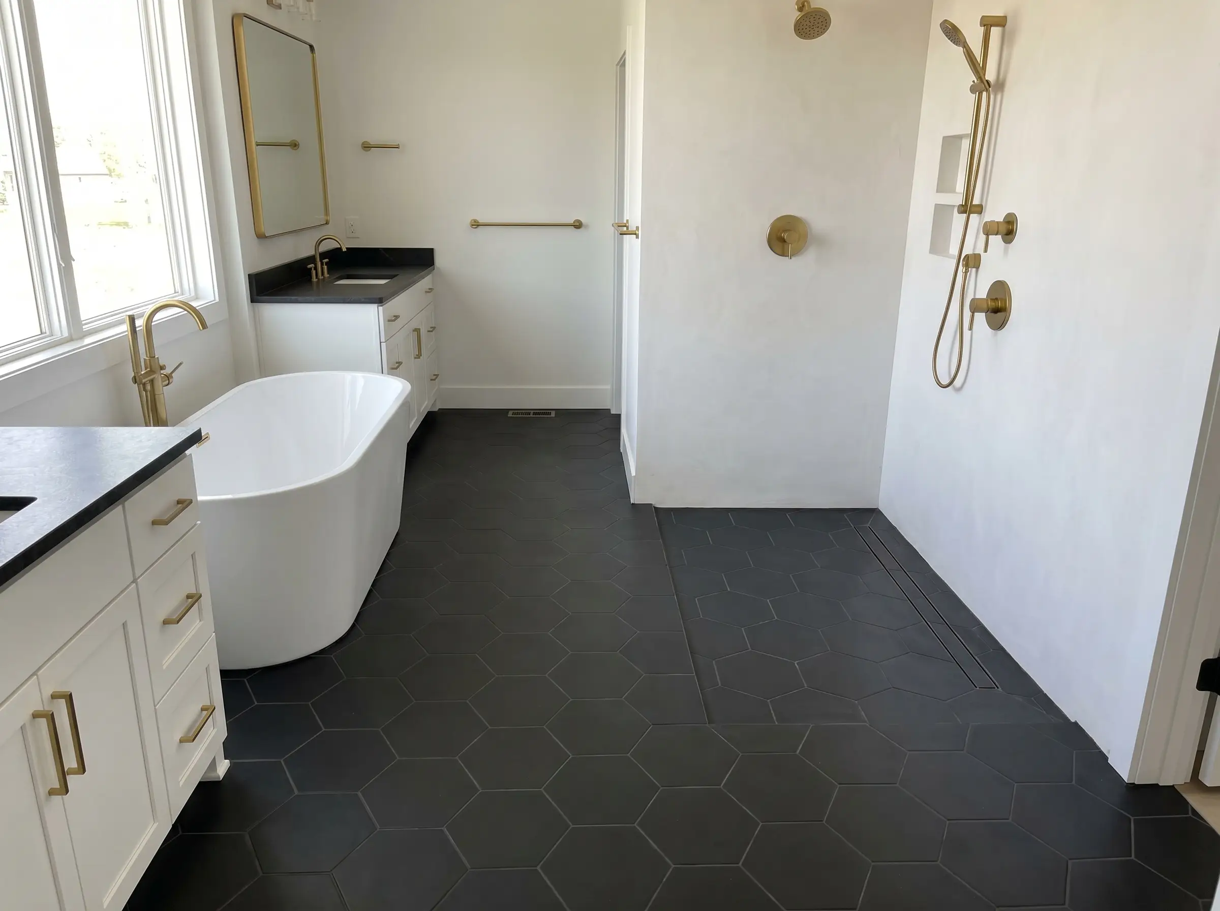 A high-contrast white and gold bathroom featuring a white freestanding tub and sharp gold hardware anchored by matte black porcelain hexagon floor tiles.