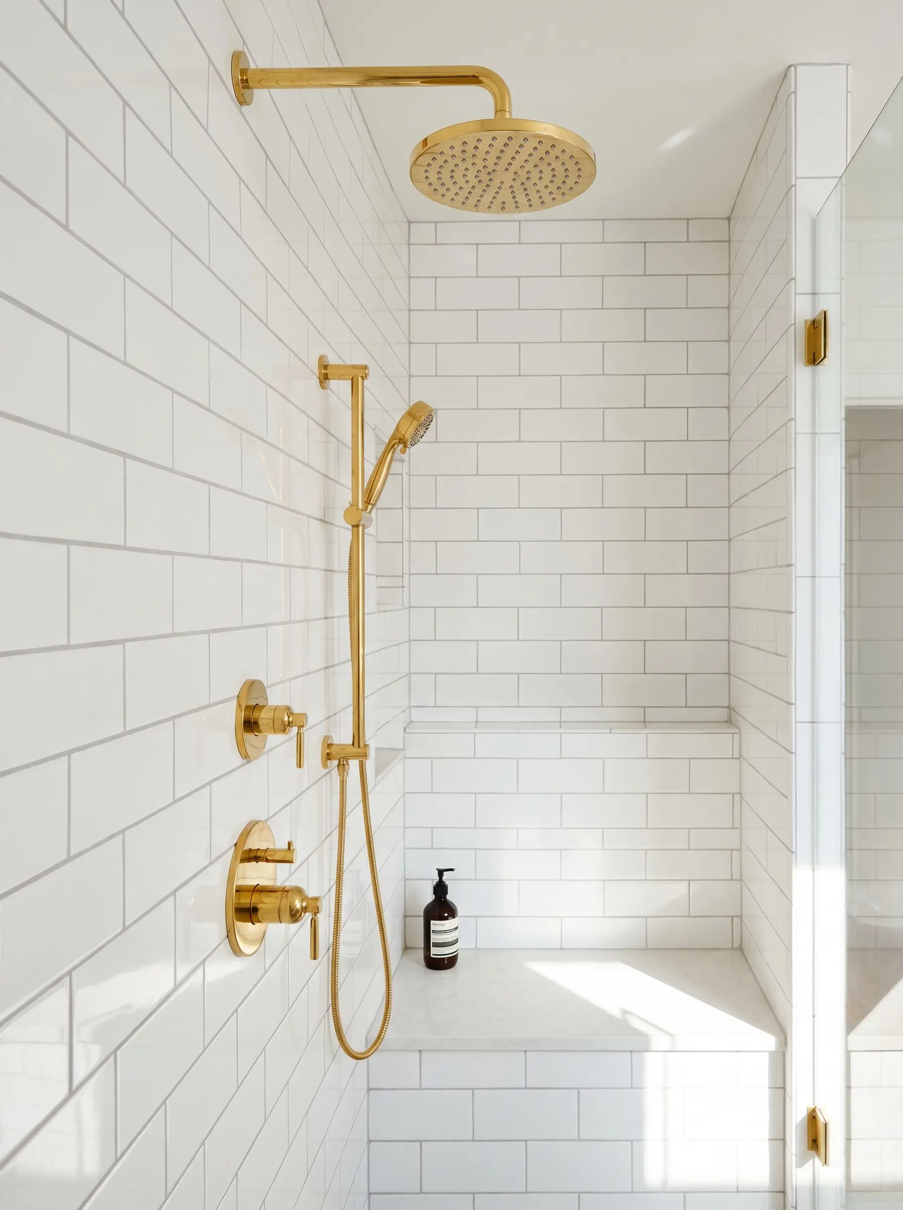 Bright gold shower hardware is mounted on a wall of ceramic white subway tiles defined by warm pale gray sanded grout.